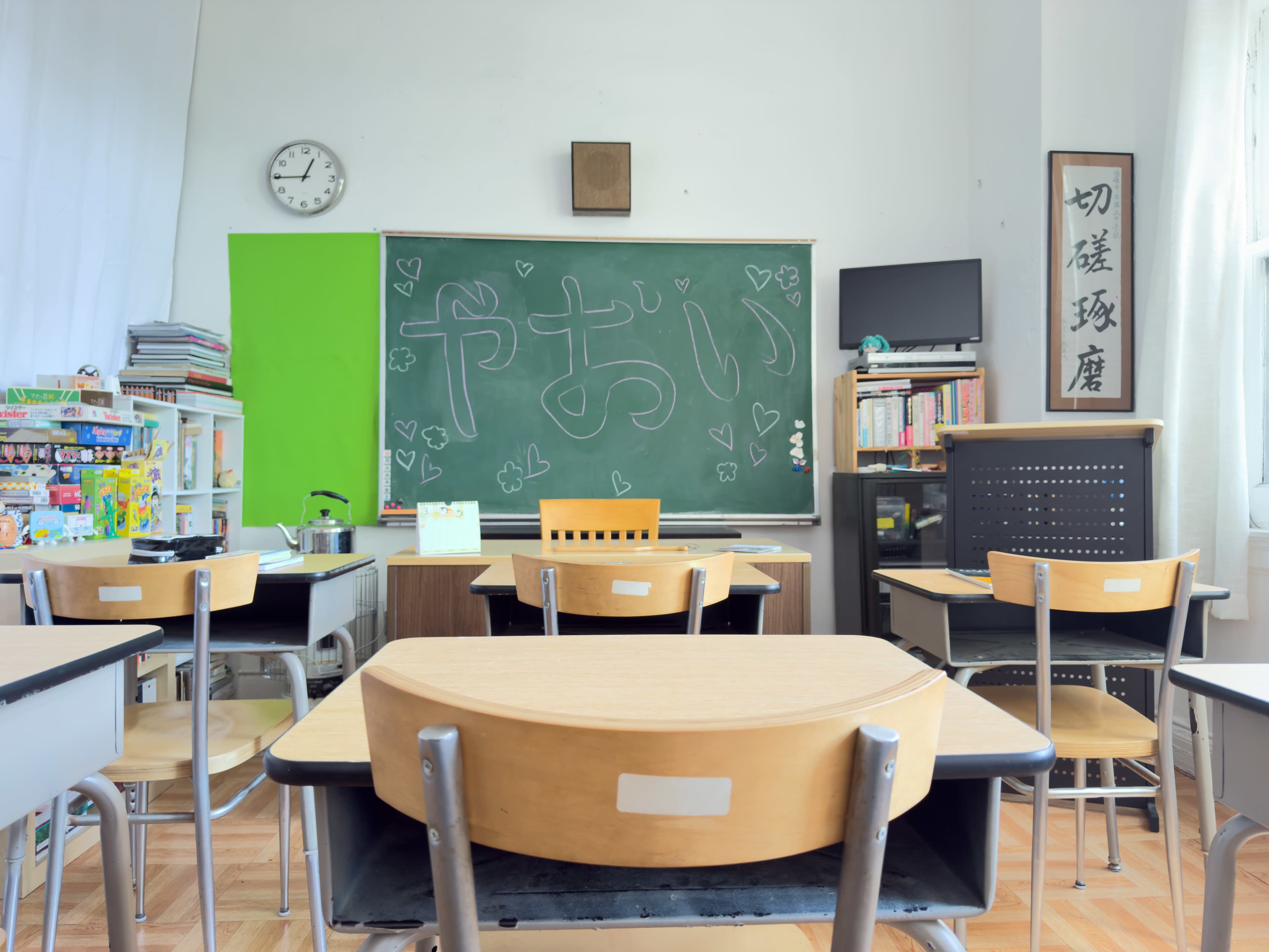 A classroom is set up with wooden desks and chairs facing a chalkboard, which features handwritten characters and drawings.