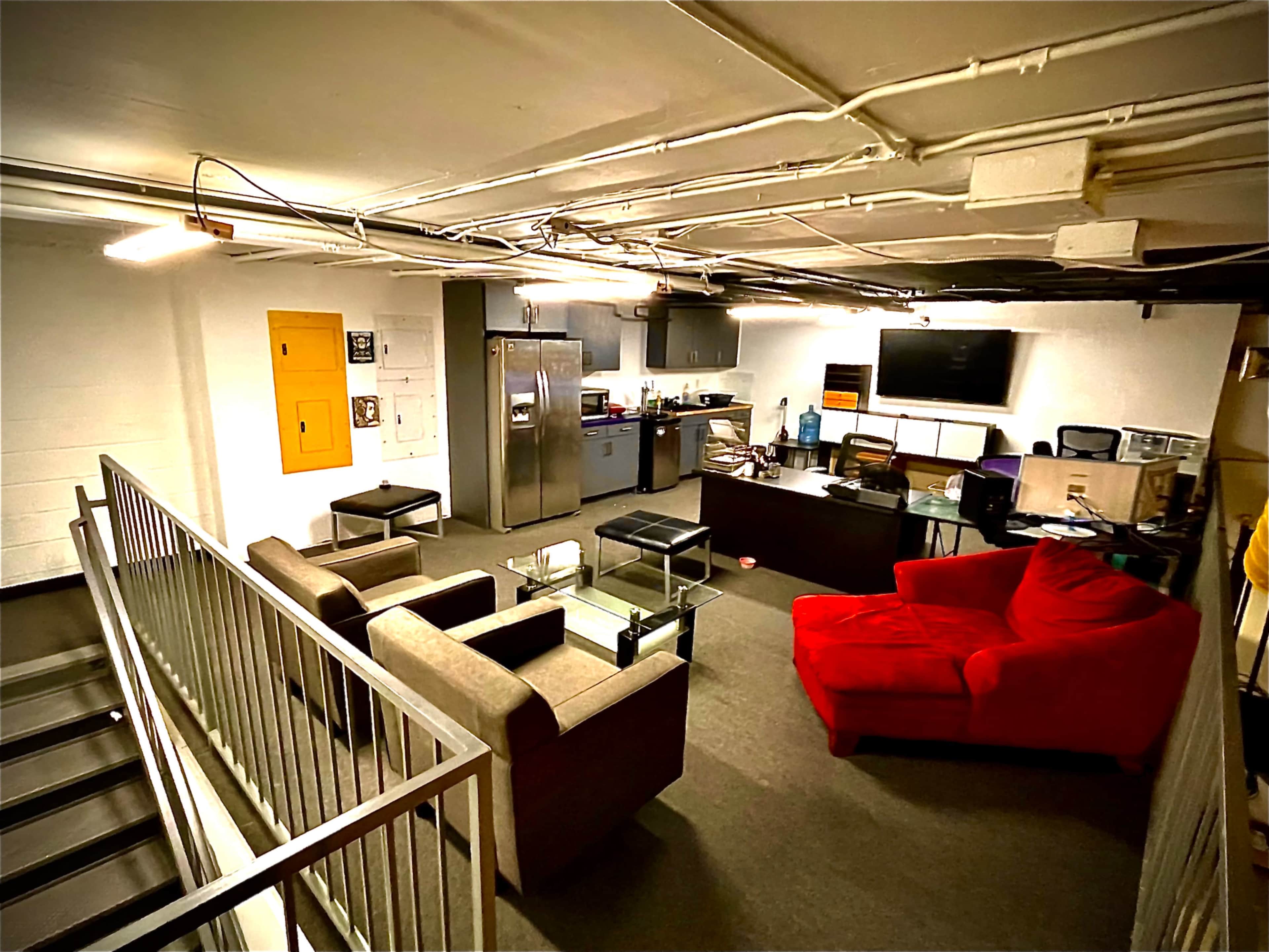 The image shows a basement area furnished with a red couch, gray chairs, a kitchen space with stainless steel appliances, and a desk with a computer monitor.