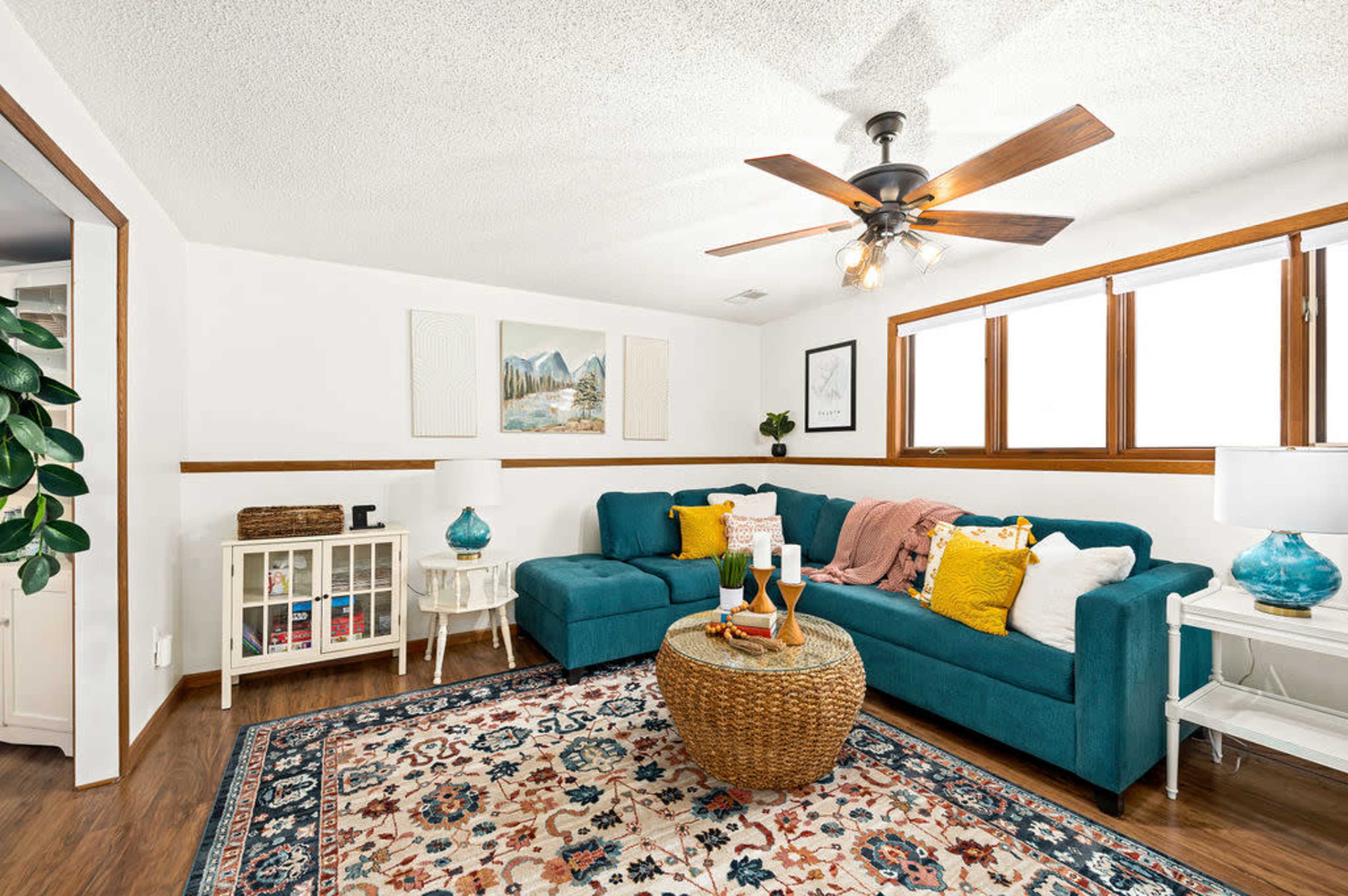 The image showcases a cozy living room featuring a teal sectional sofa, a decorative area rug, and large windows that allow natural light to fill the space.