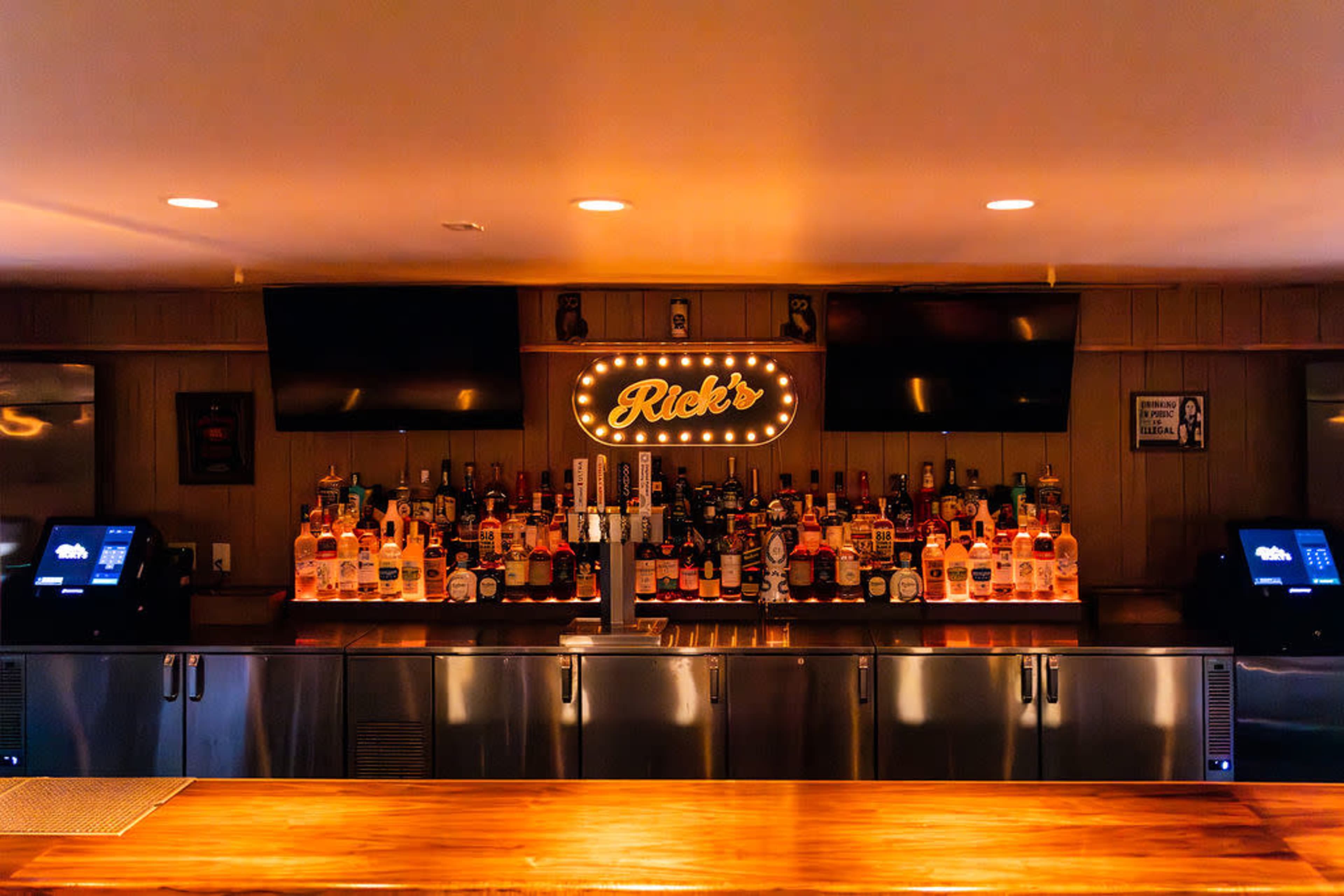 The image shows a brightly lit bar with a variety of liquor bottles arranged on shelves behind a wooden counter.