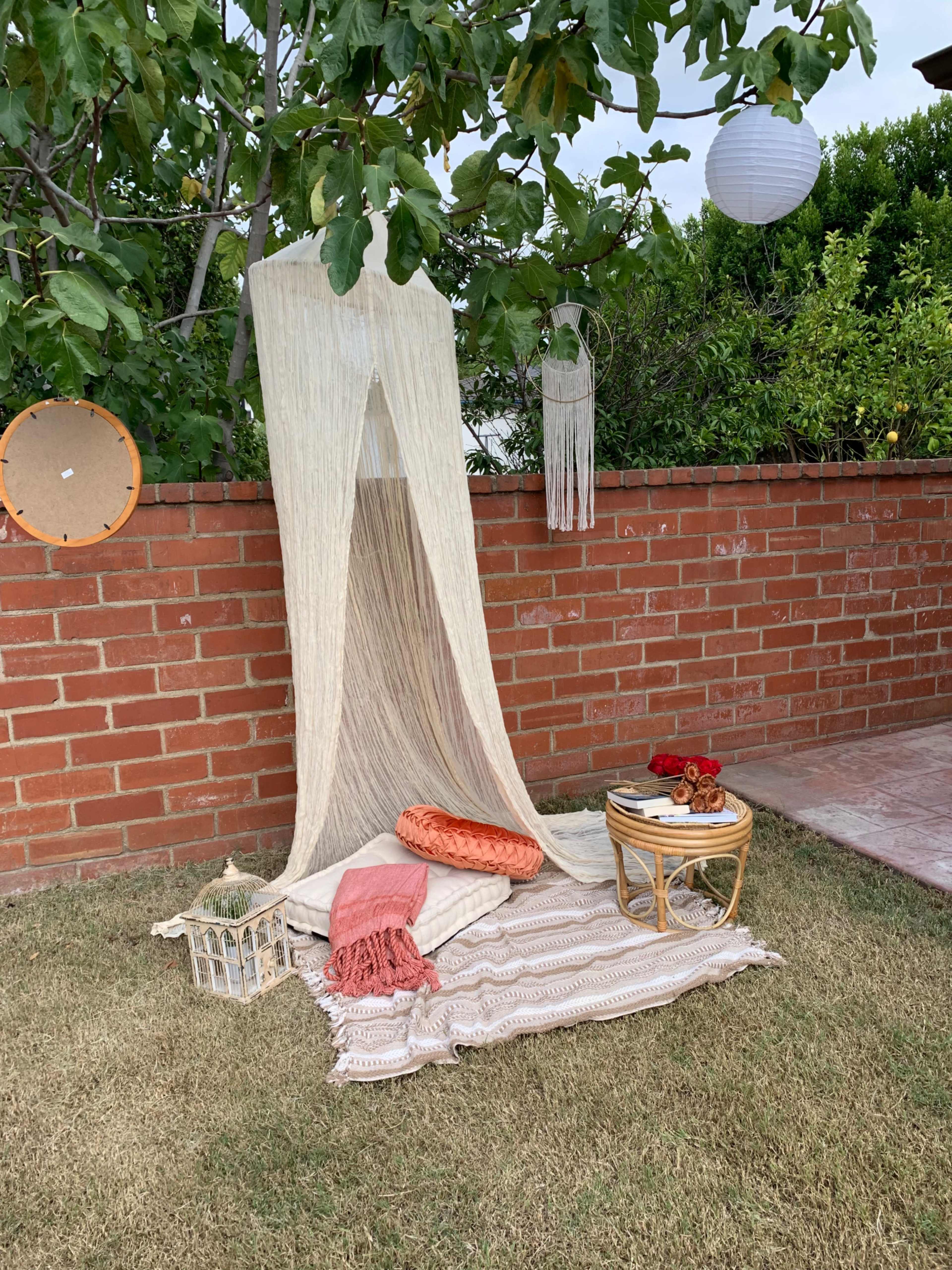 A cozy outdoor seating area features a draped fabric backdrop, a decorative rug, cushions, and a small round table with snacks arranged in front of a brick wall.