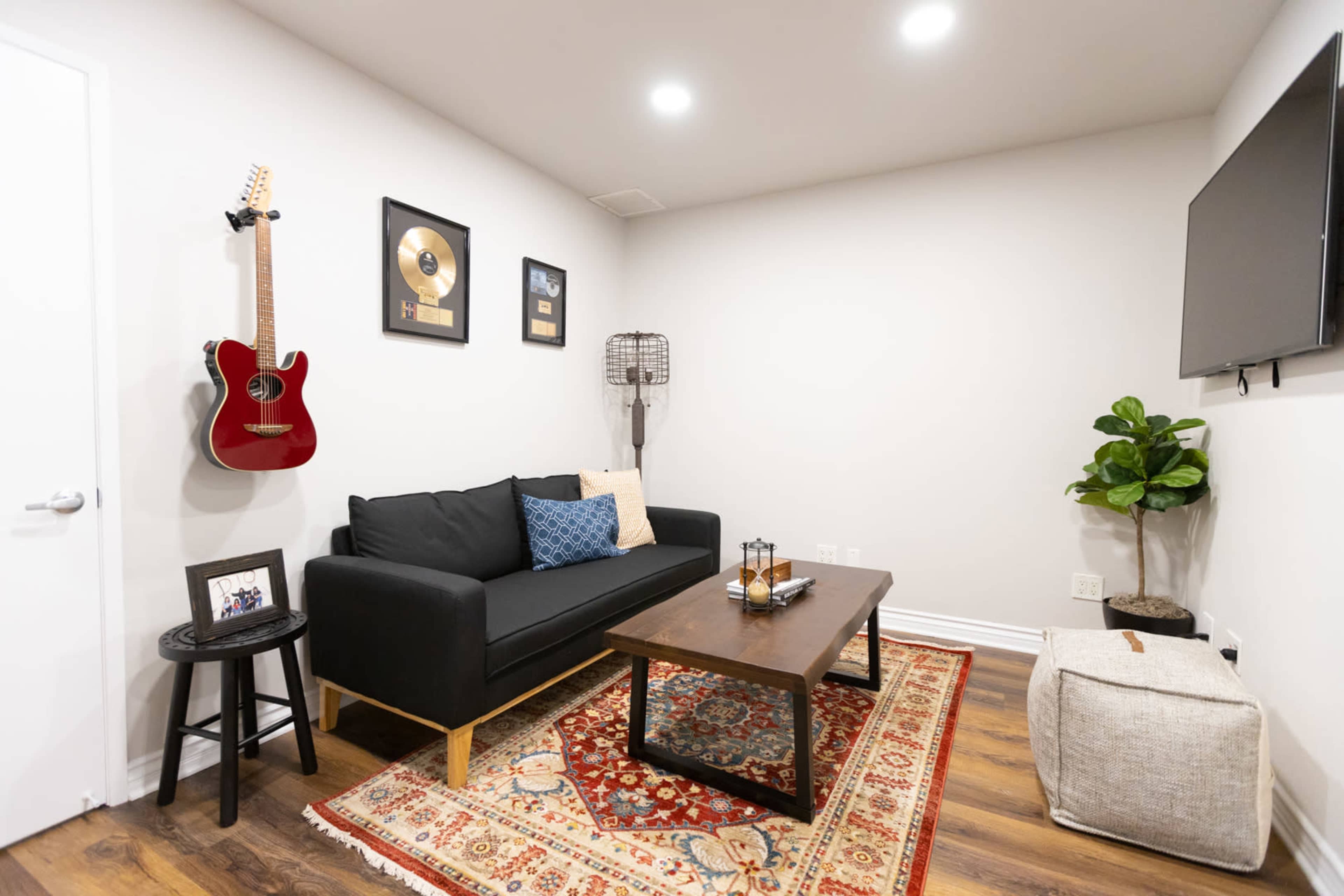The image shows a cozy living room with a black couch, a wooden coffee table, a guitar mounted on the wall, and a plant in the corner.
