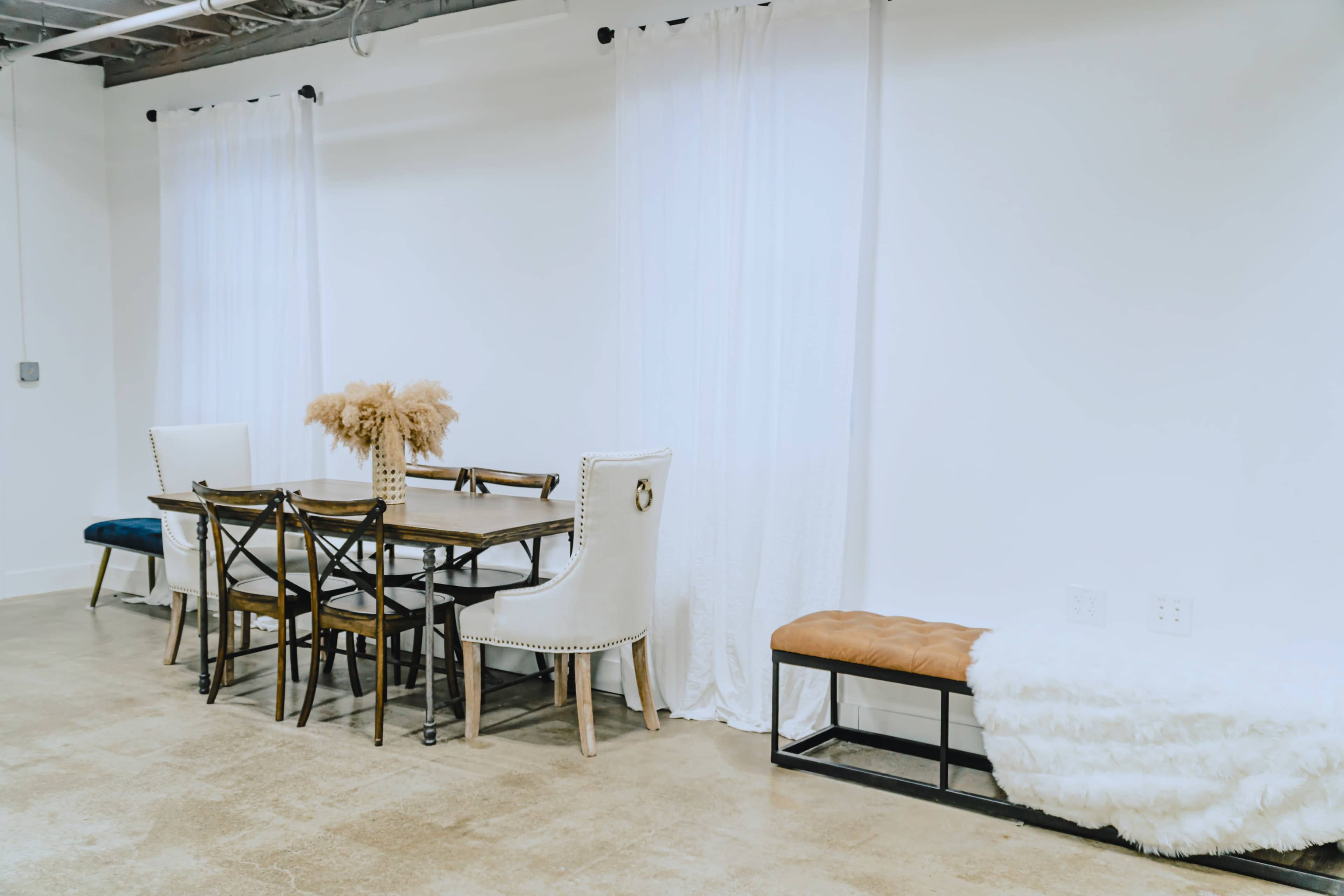 A dining area features a wooden table surrounded by chairs, with a bench and decorative plants against a white wall.