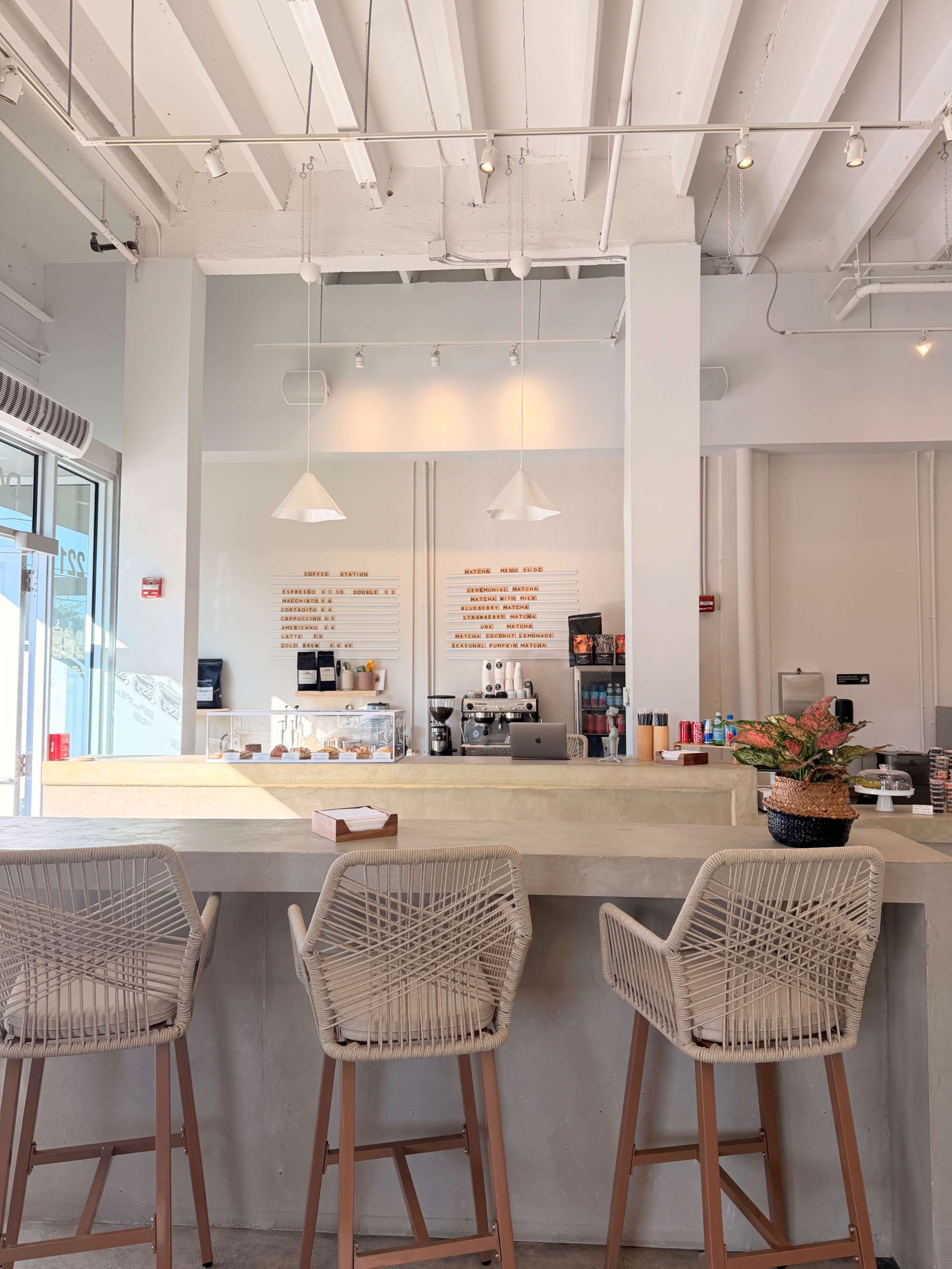 The image shows a modern café interior with a light-filled space, featuring a counter with coffee-making equipment, a menu on the wall, and several barstools.