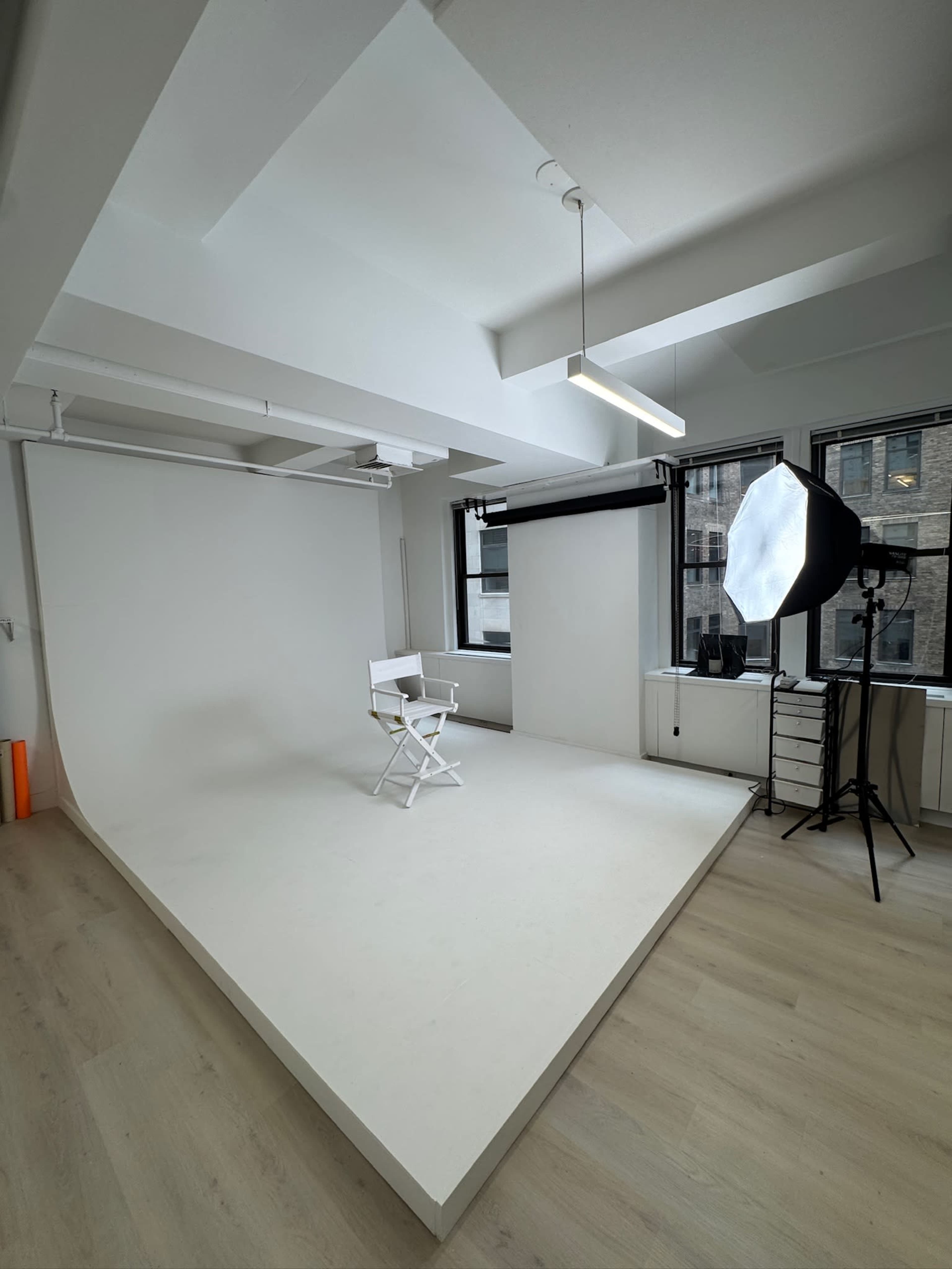 The image shows a modern photography studio with a white backdrop, a director's chair, and lighting equipment set up near large windows.