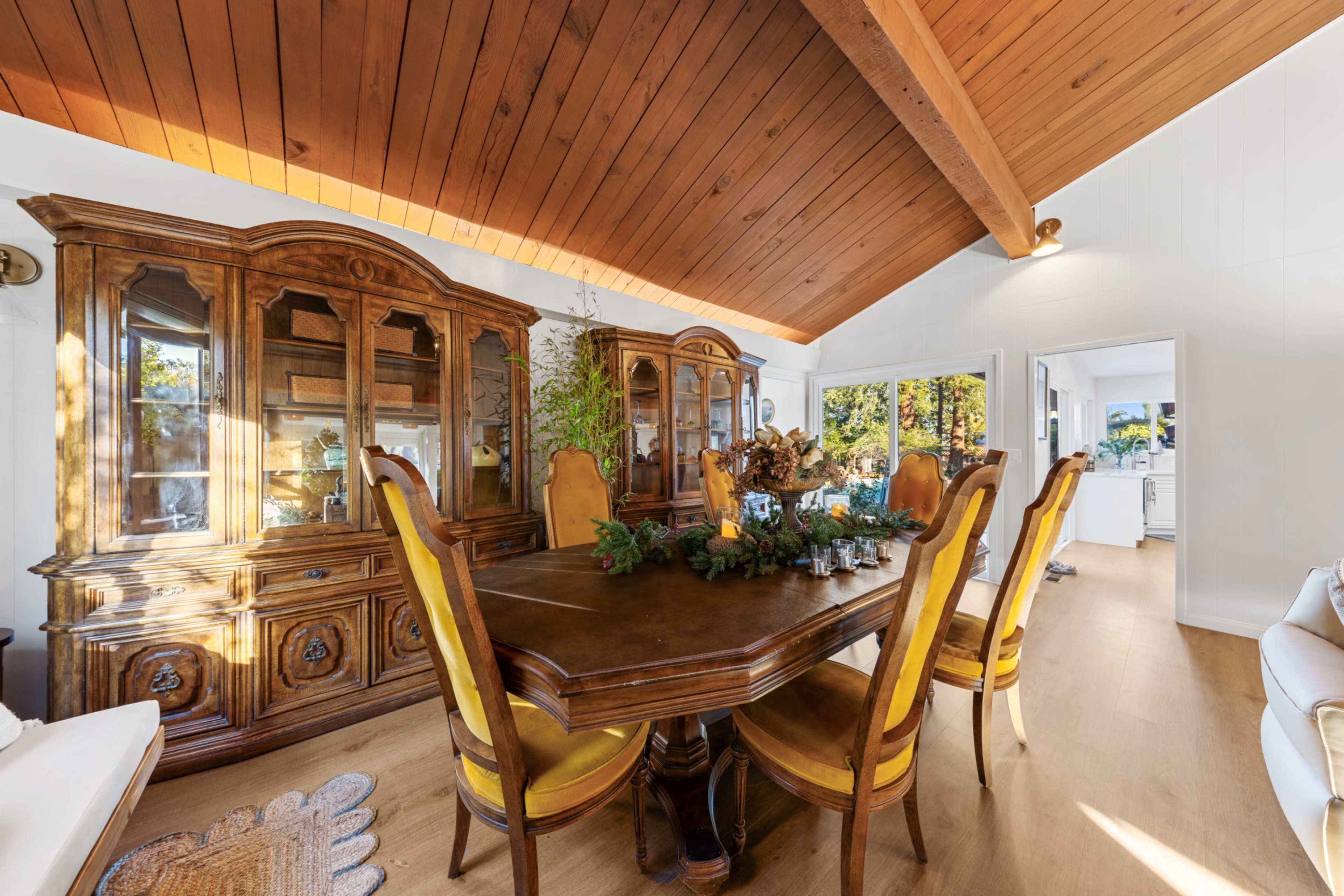 The image shows a dining room with a wooden table surrounded by yellow upholstered chairs and a large antique cabinet against the wall.