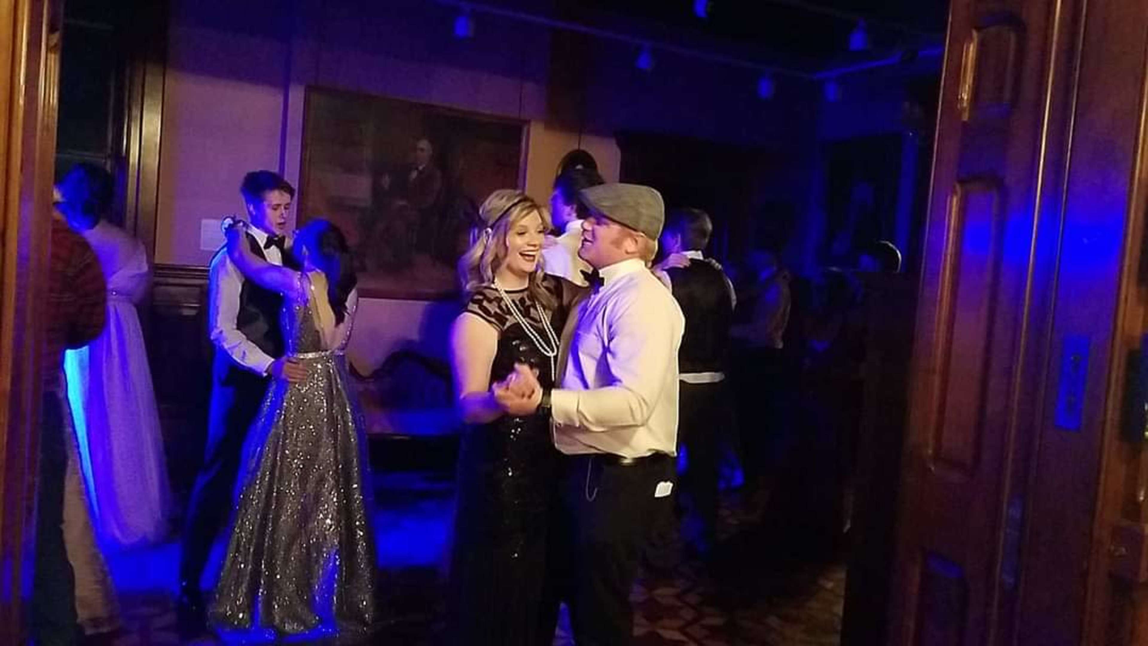 A couple dances closely in a lit room filled with guests, with some wearing formal attire and others in casual clothing.