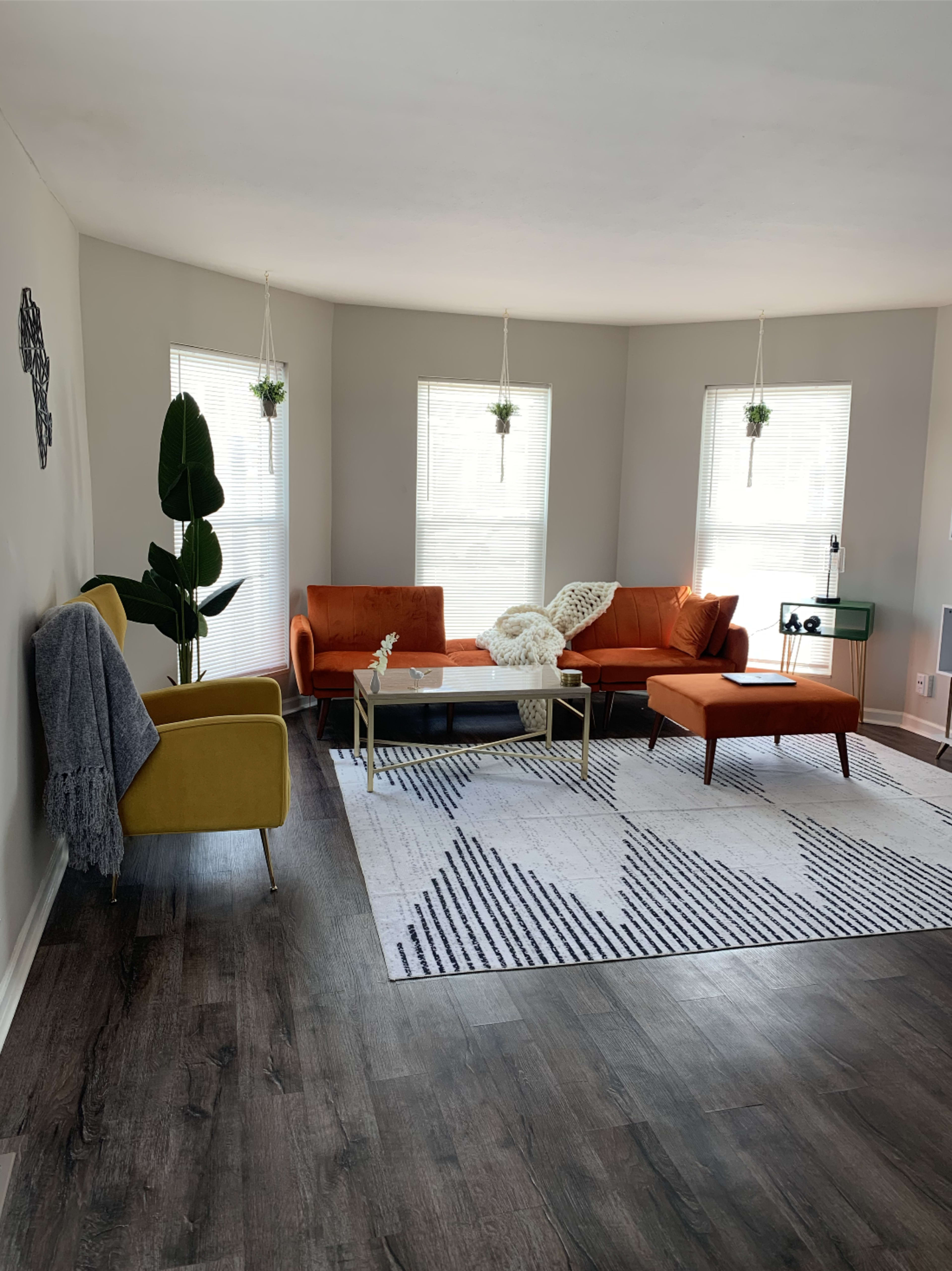 The living room features three colorful sofas, a coffee table, and large windows with plants hanging above.