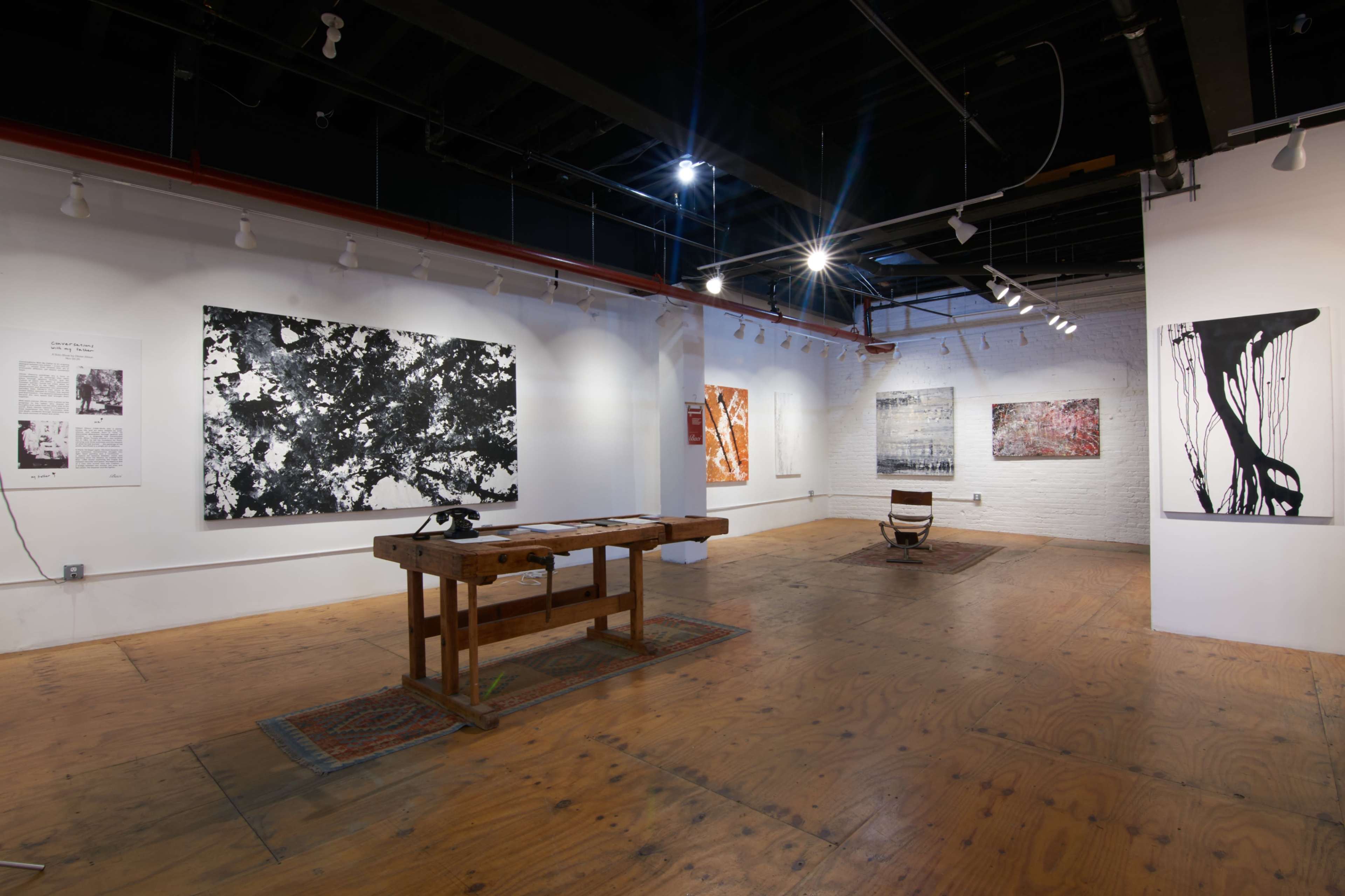 The image shows an art gallery with white walls, showcasing several large abstract paintings, and a wooden table in the center of the room.