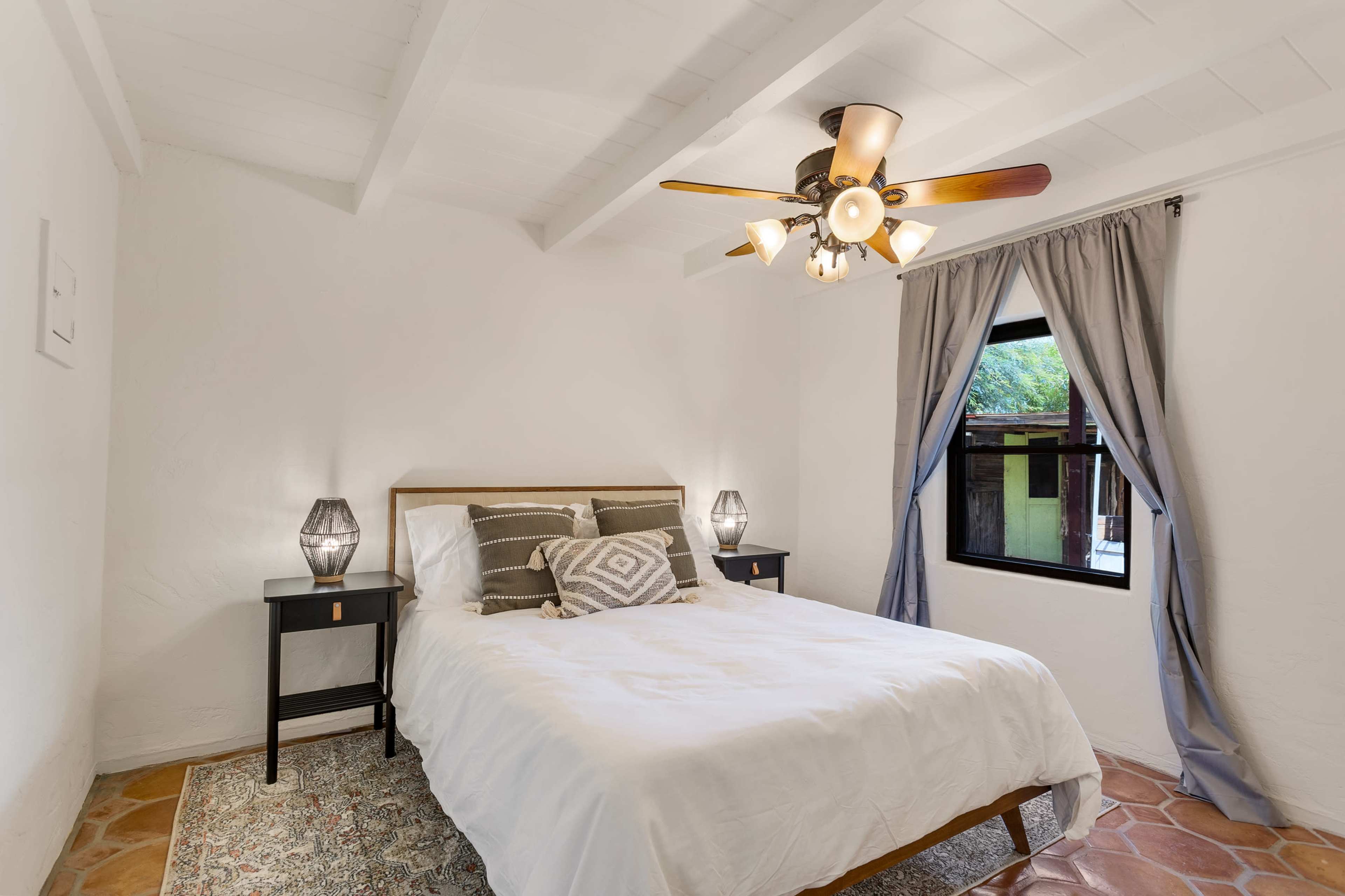 A well-lit bedroom featuring a bed with decorative pillows, two nightstands with lamps, and a window with curtains.