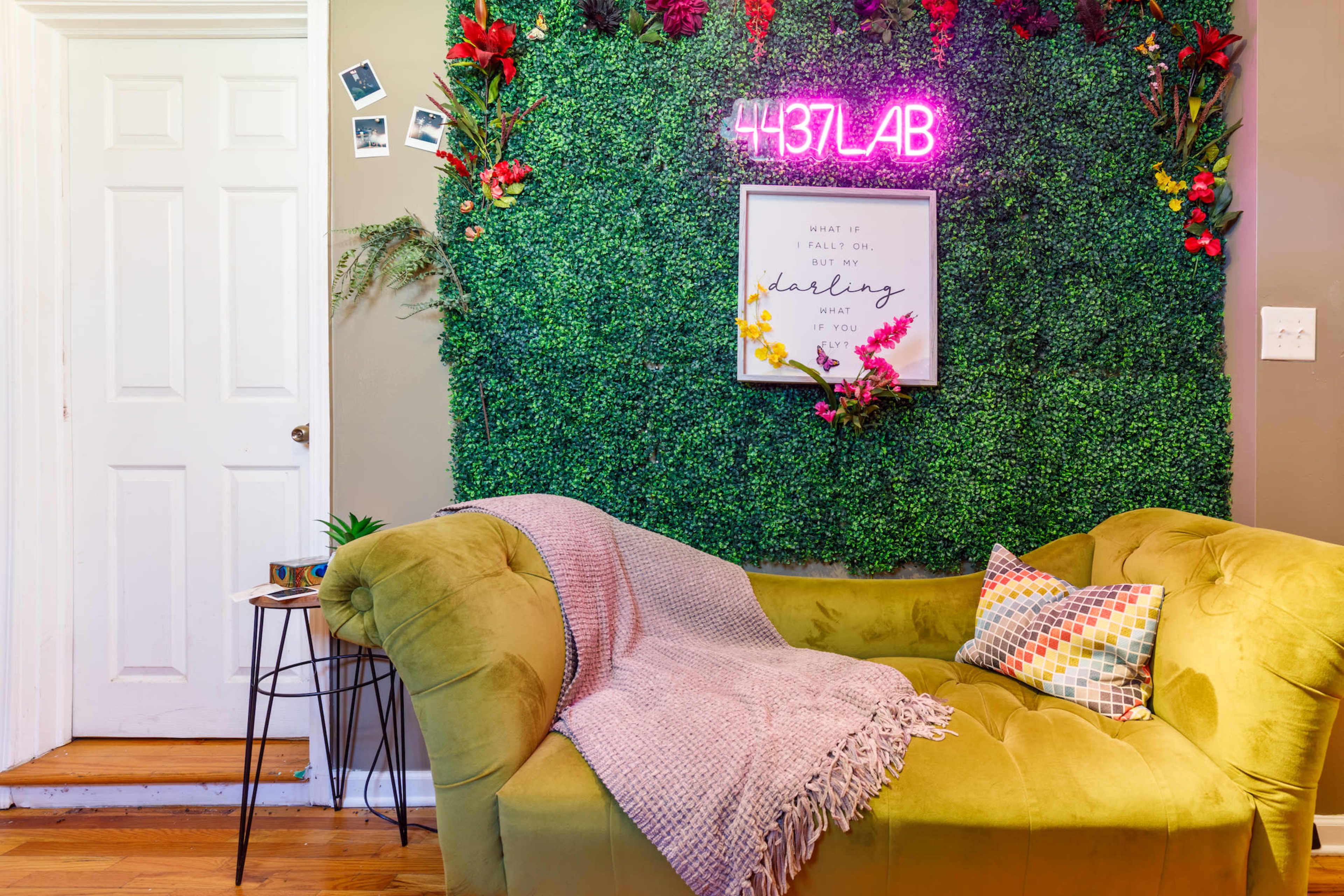 A cozy seating area features a green backdrop with a neon sign, surrounded by plants and decorative elements.
