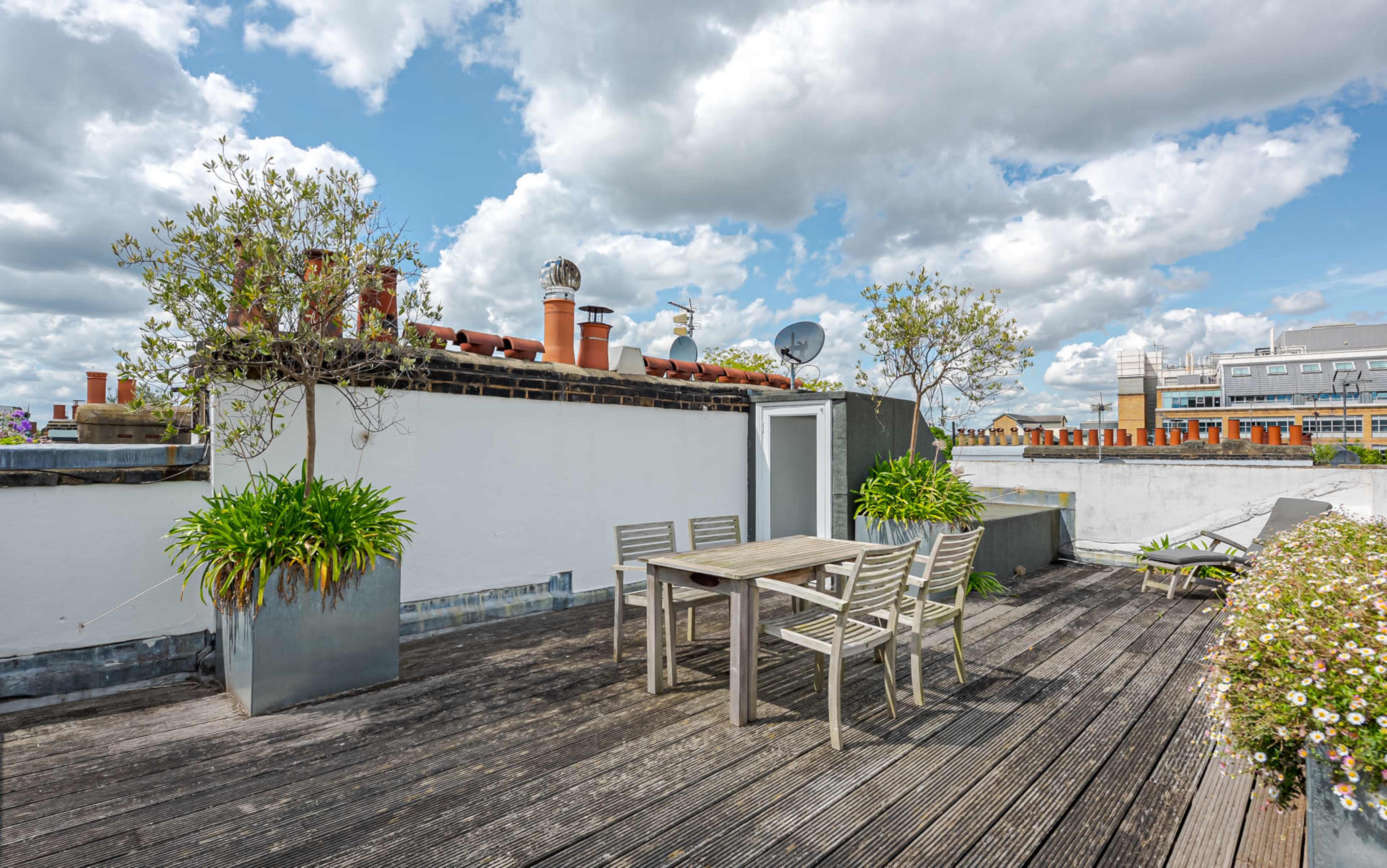 The image shows a rooftop terrace with a wooden deck, a metal table and chairs, planters with greenery, and a backdrop of clouds and nearby buildings.