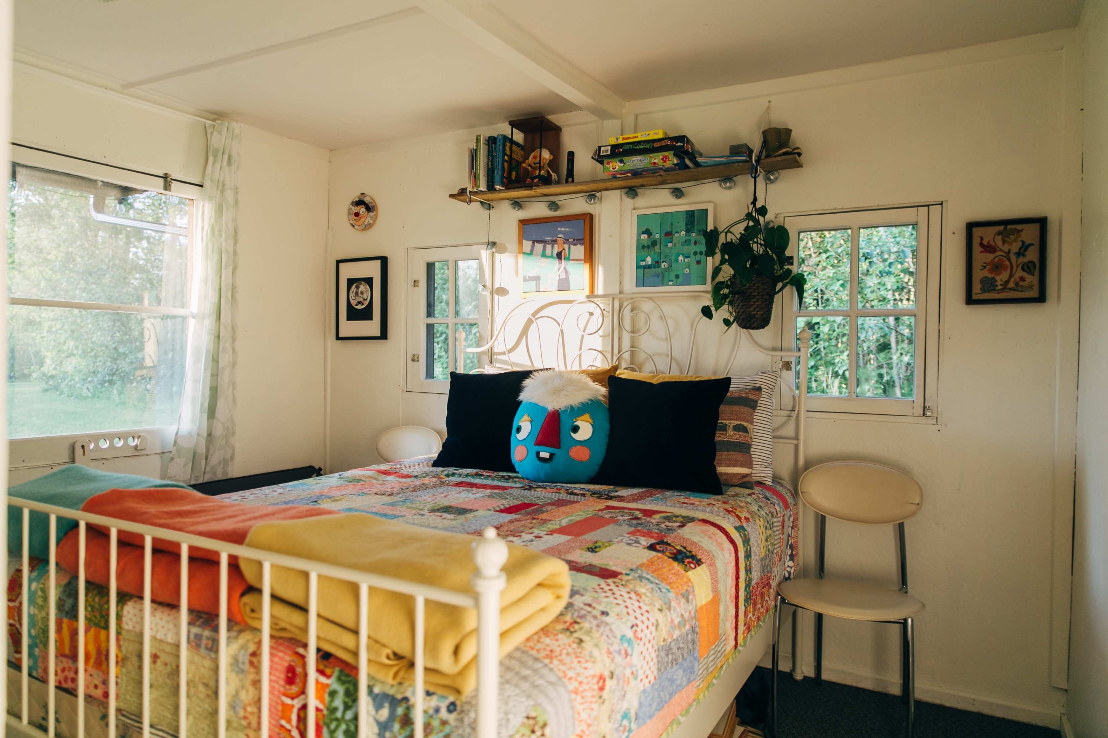 A cozy bedroom featuring a quilt-covered bed, a colorful pillow with a cartoon face, and shelves filled with books and plants.