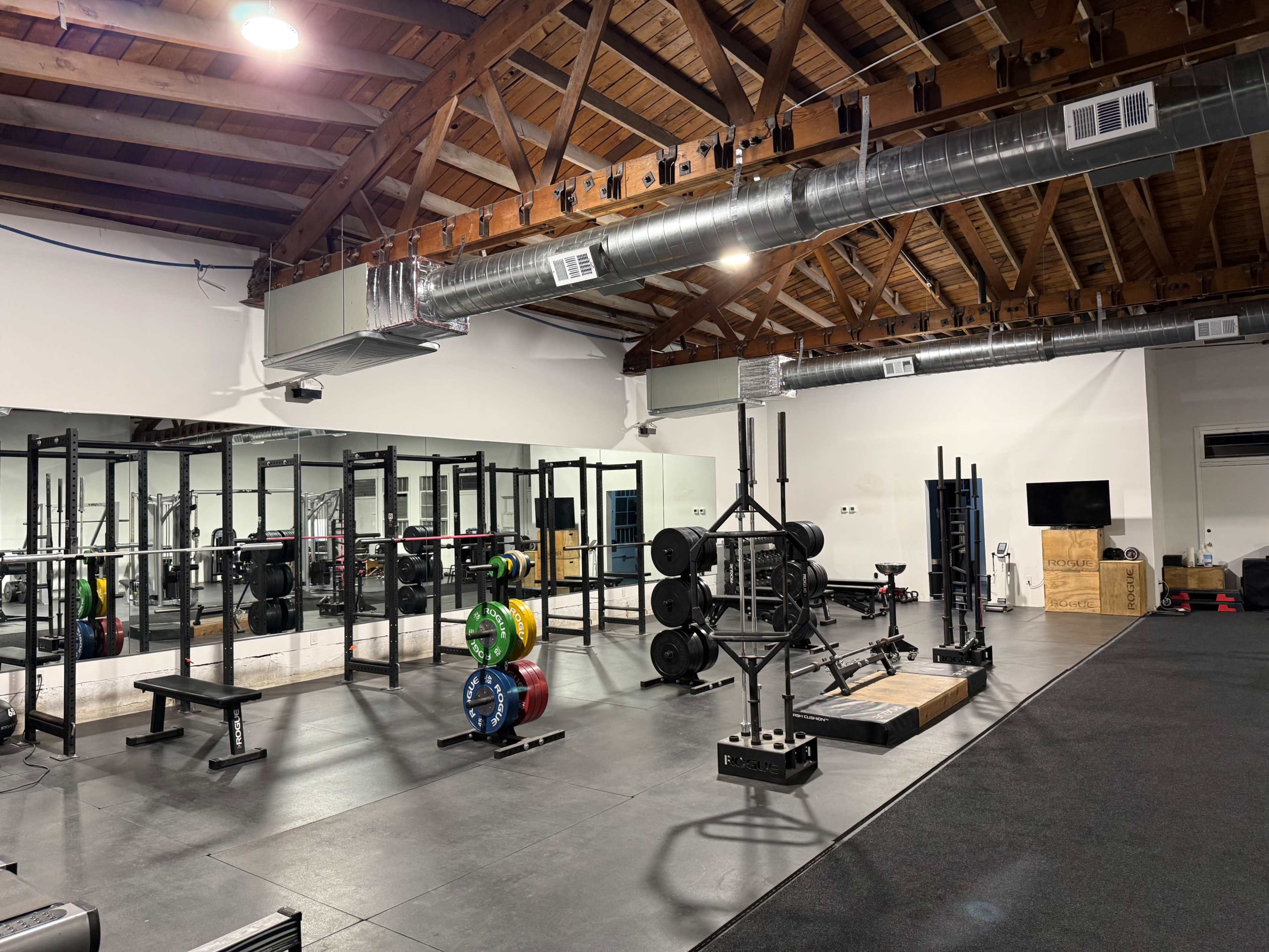 The image shows an indoor gym with various weightlifting equipment, including benches, barbells, and weight plates, set in a spacious room with wooden beams and mirrored walls.