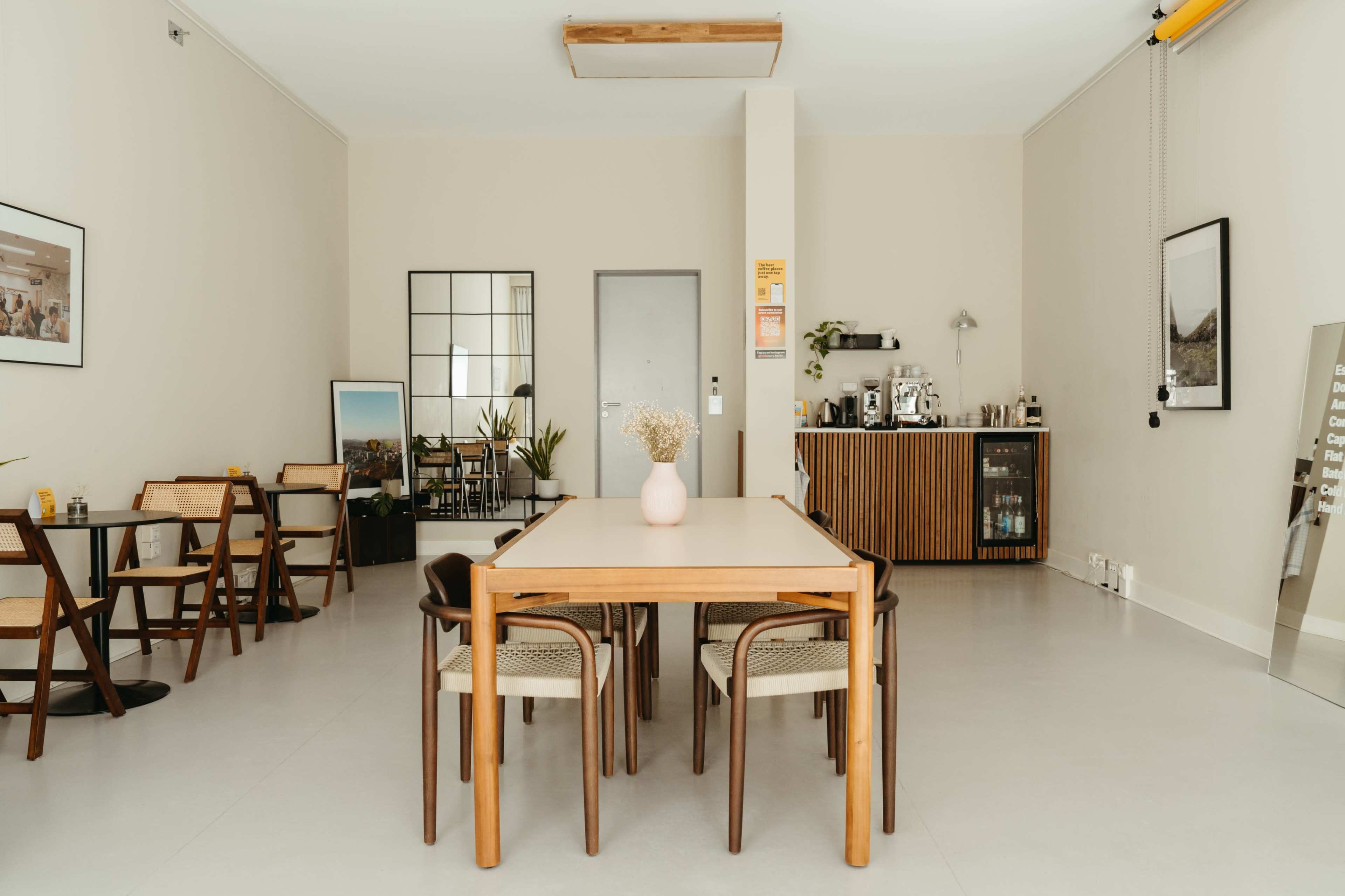 A spacious, minimalist interior features a central wooden dining table surrounded by chairs, with a mirrored wall and a side cabinet visible in the background.
