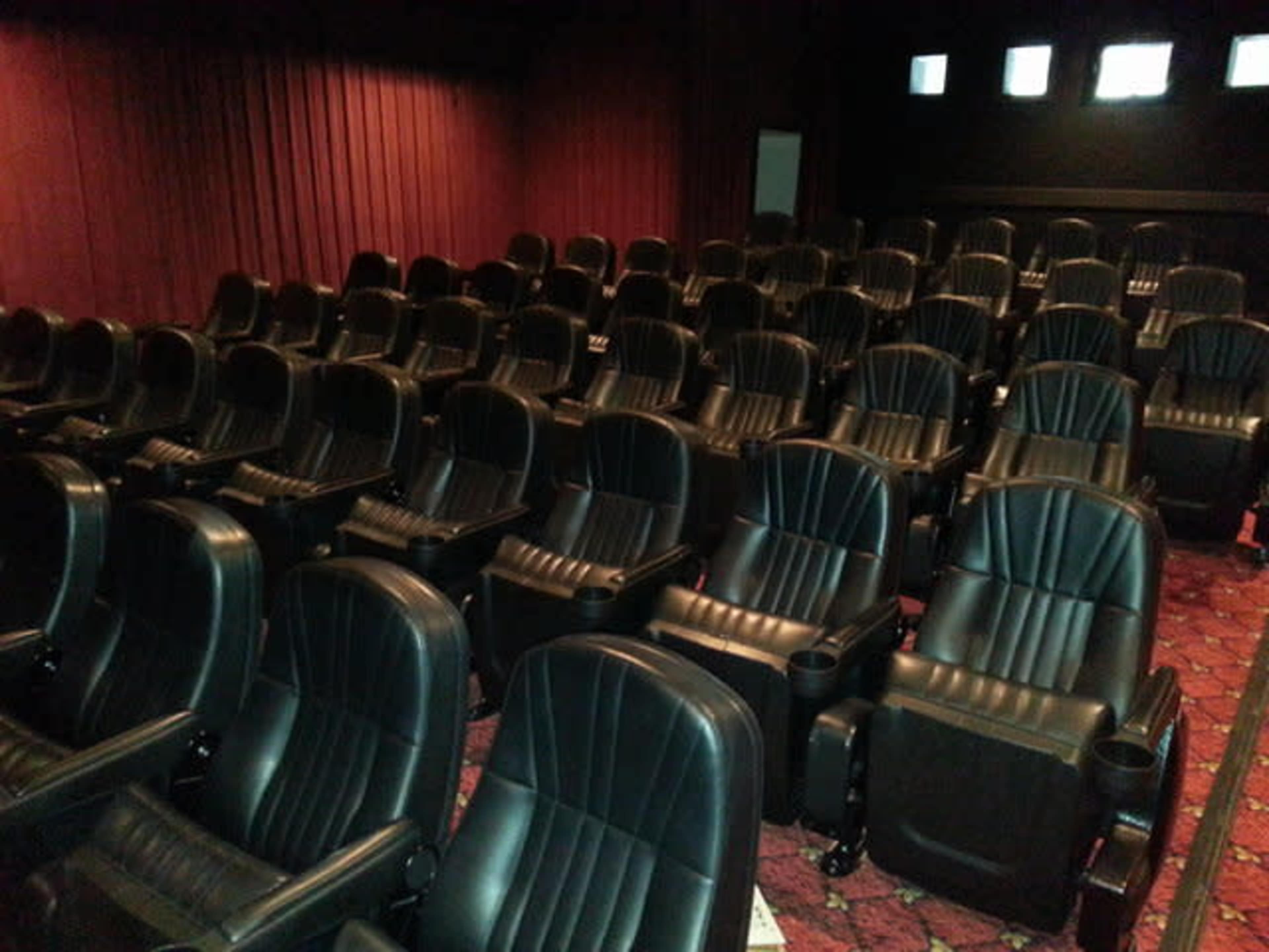 The image shows a movie theater with rows of empty black leather recliner seats against a red curtain backdrop.
