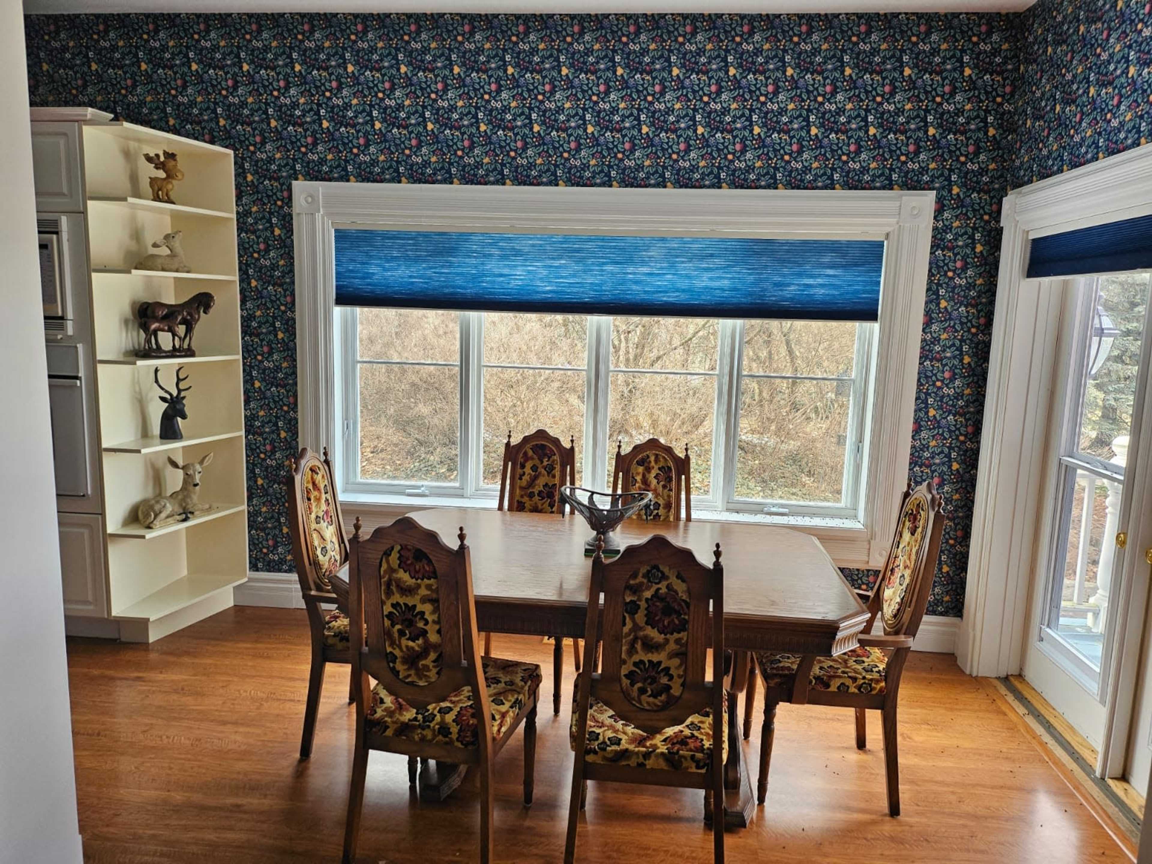 A dining room features a wooden table surrounded by ornate chairs, with large windows offering a view of a landscape beyond a floral wallpapered wall.