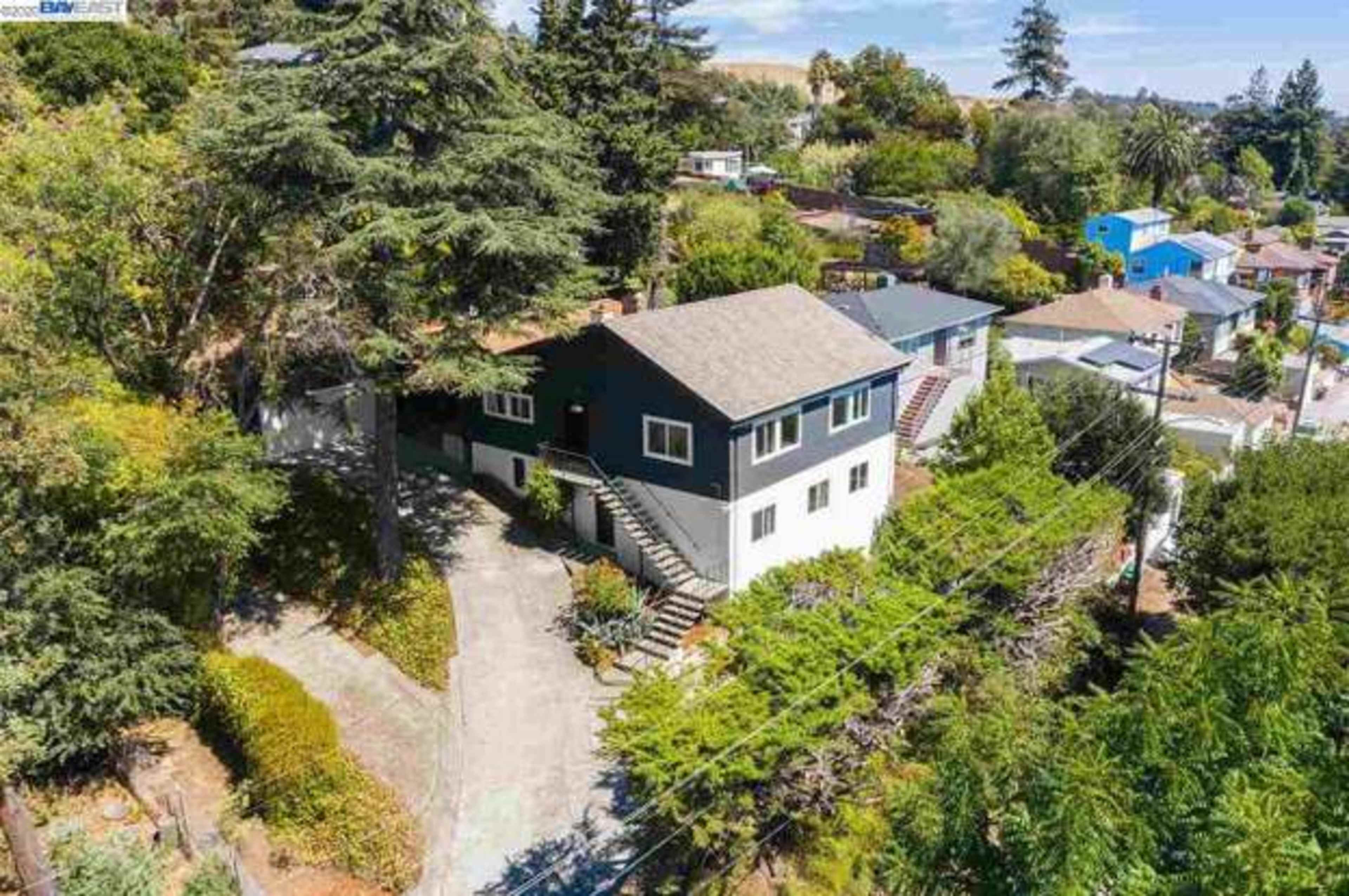 A two-story house situated on a hillside with surrounding trees and a winding driveway.