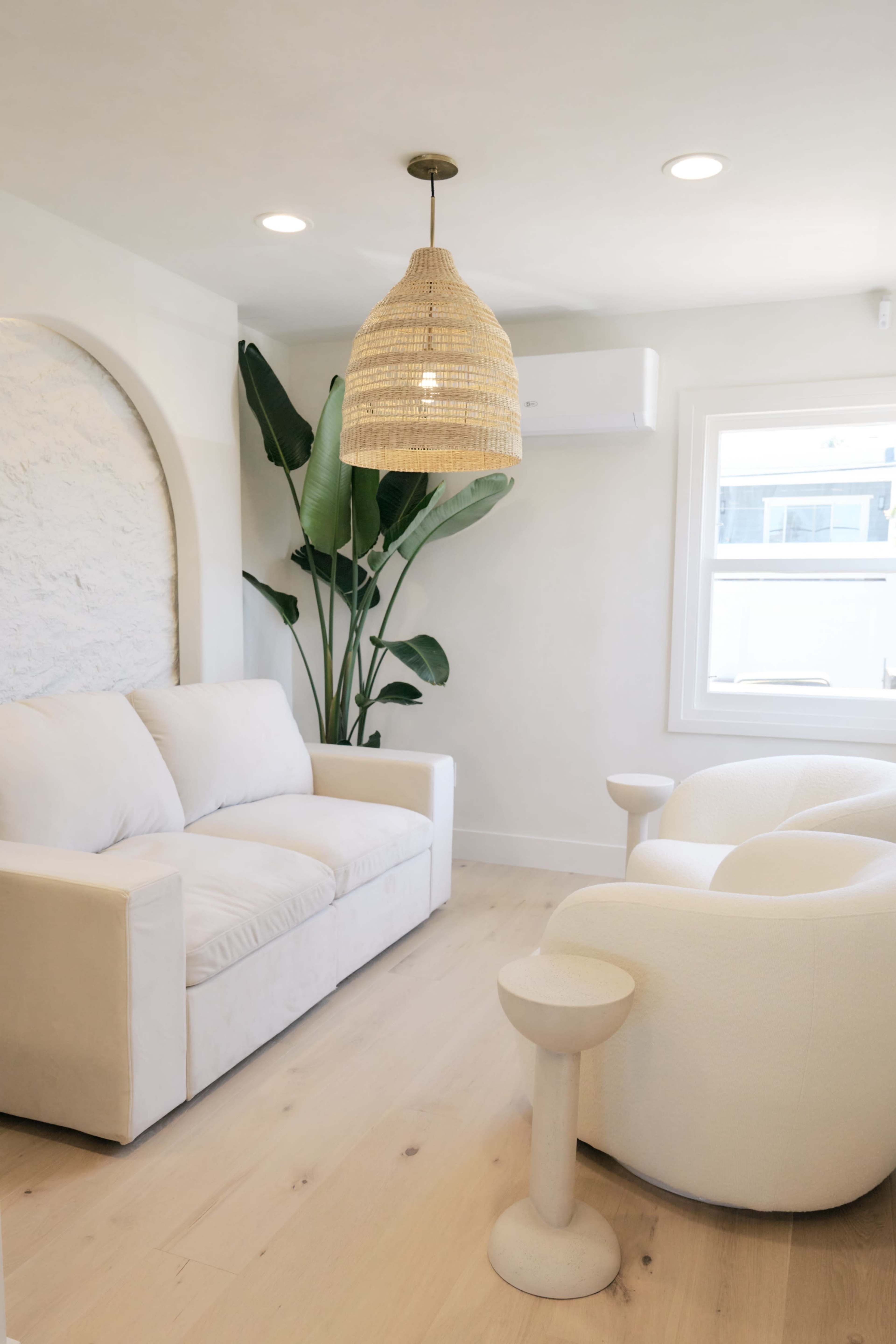 The image shows a modern living room with a white sofa, a sculptural white chair, and a large potted plant beside a window.