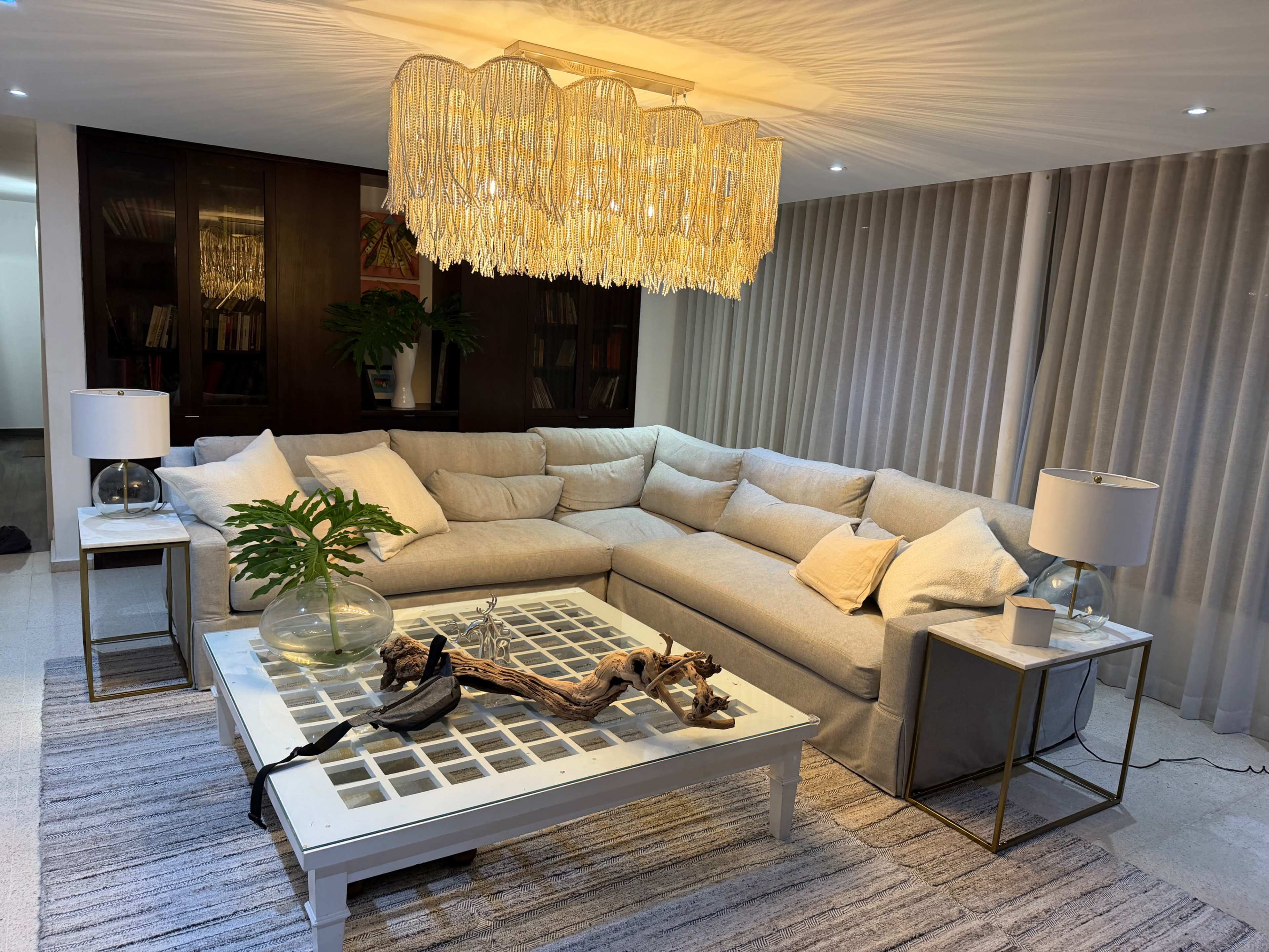 A modern living room features a large sectional sofa, a coffee table with a grid design, and a chandelier above, surrounded by curtains and decorative plants.
