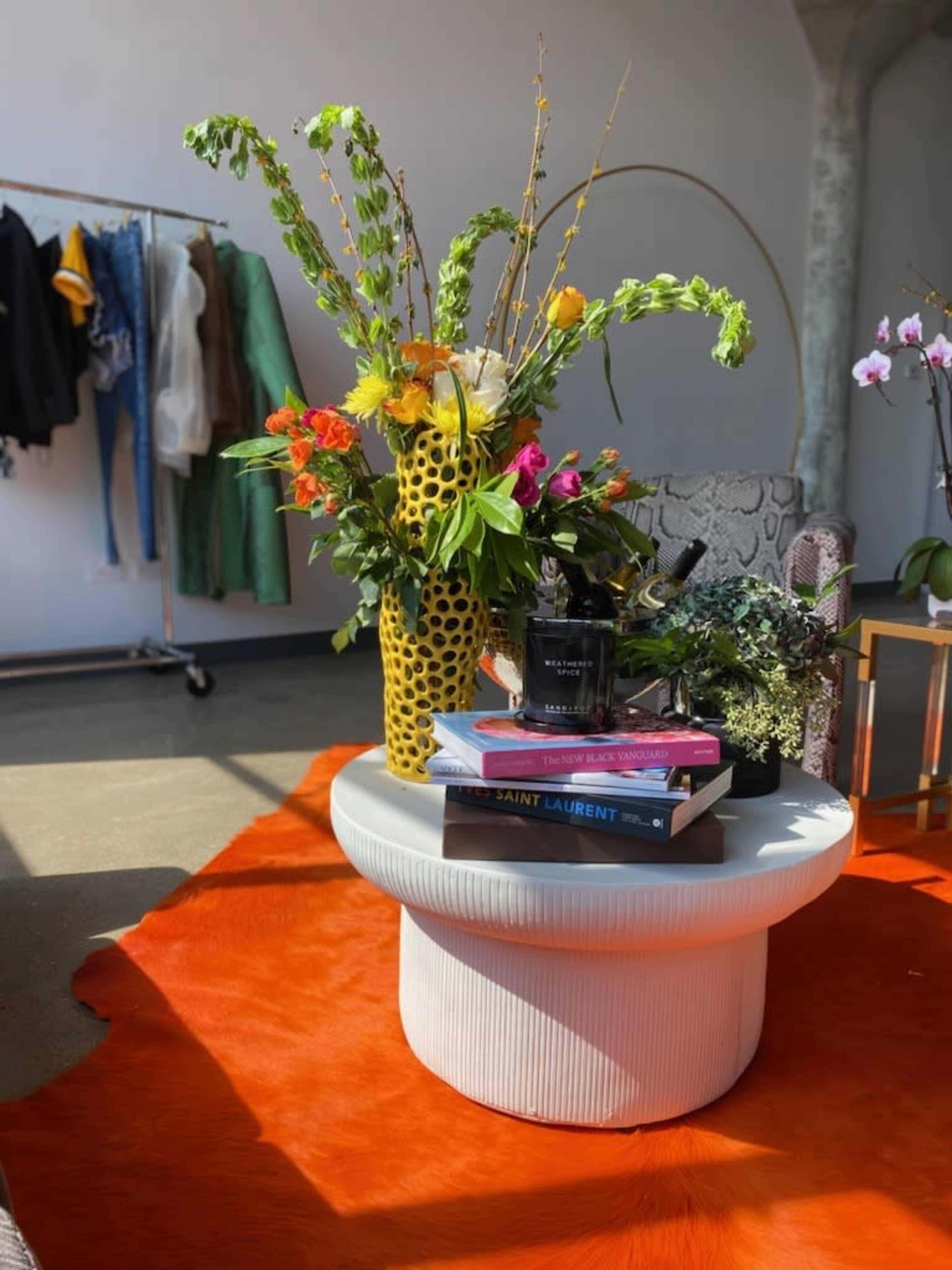 A round coffee table topped with books, a candle, and a vibrant floral arrangement sits on a bright orange rug in a styled room with clothing on a rack in the background.