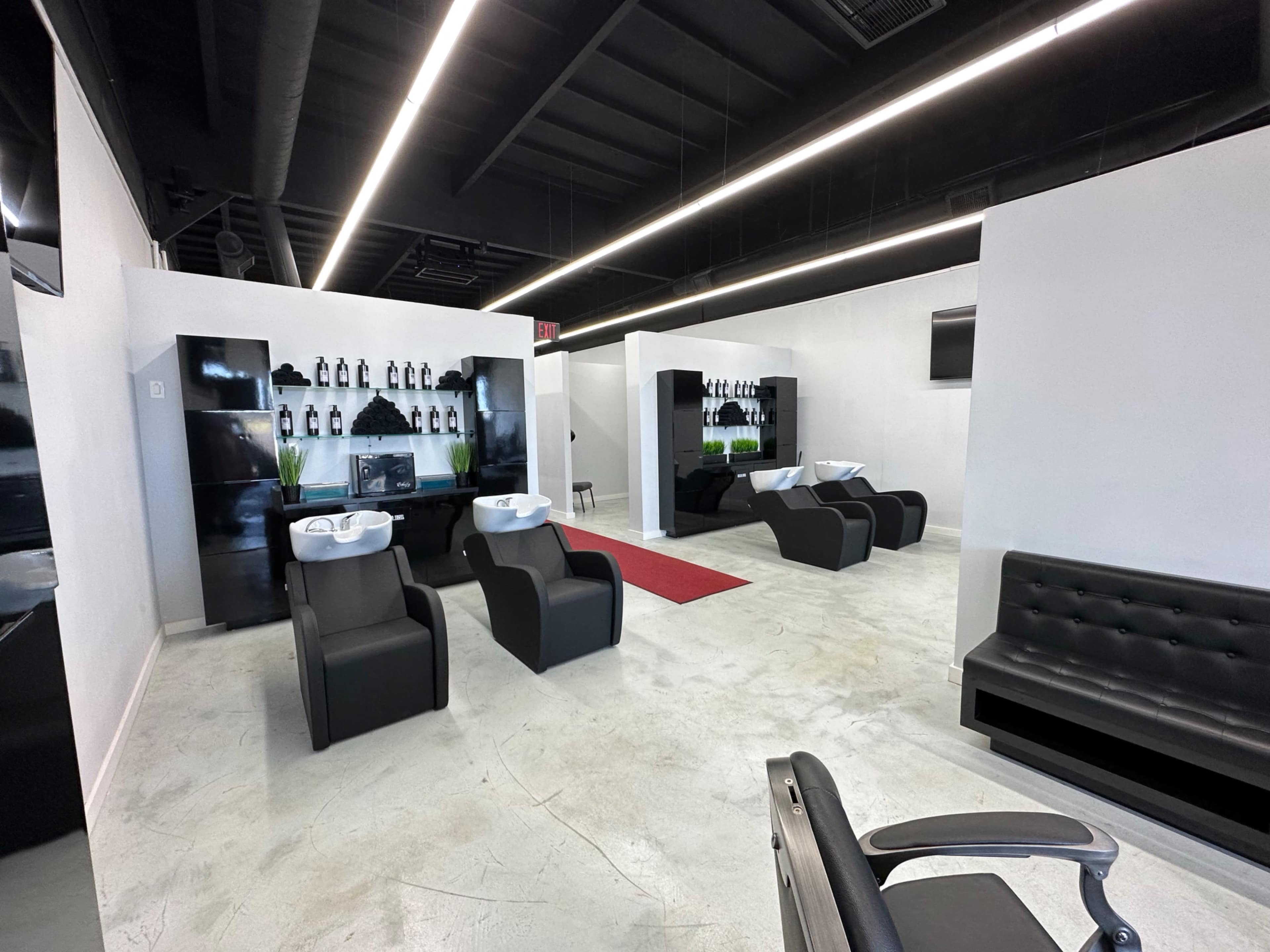 The image shows a modern hair salon featuring black styling chairs, wash stations, and a seating area with a leather couch against a minimalist white backdrop.