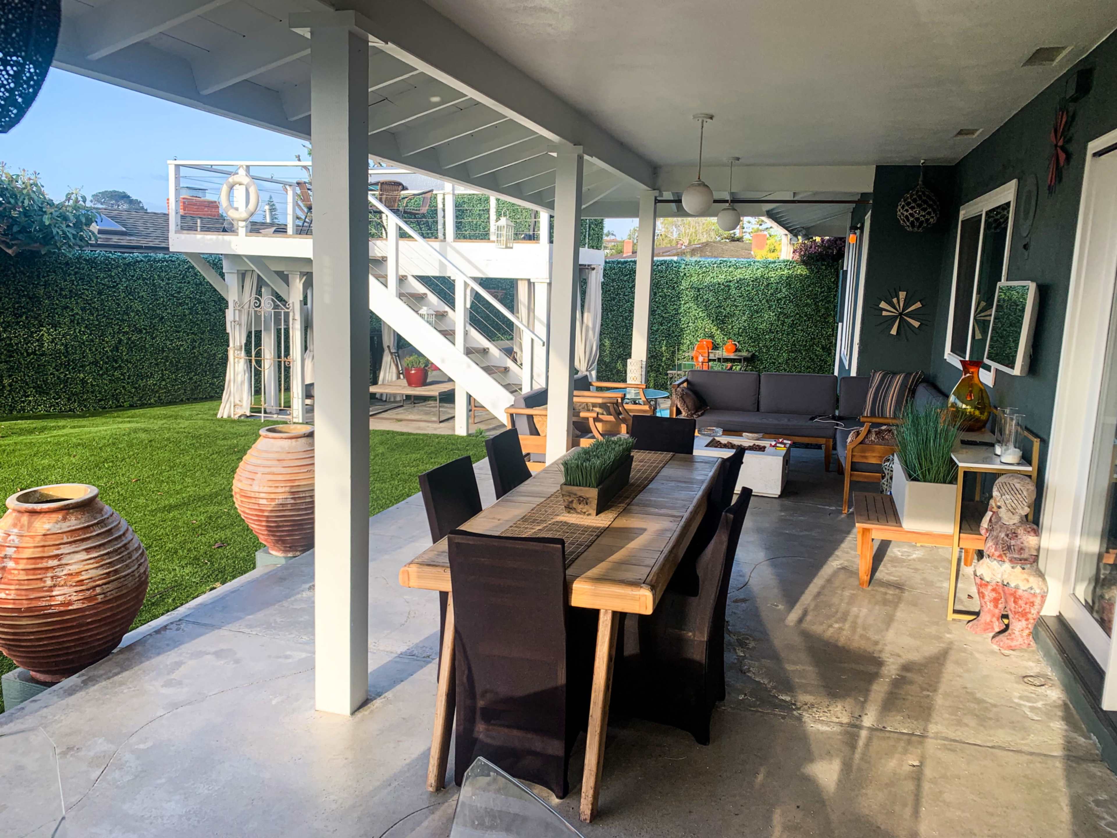 The image shows a patio area with a wooden dining table, black chairs, and outdoor furniture, surrounded by green hedges and a lawn.