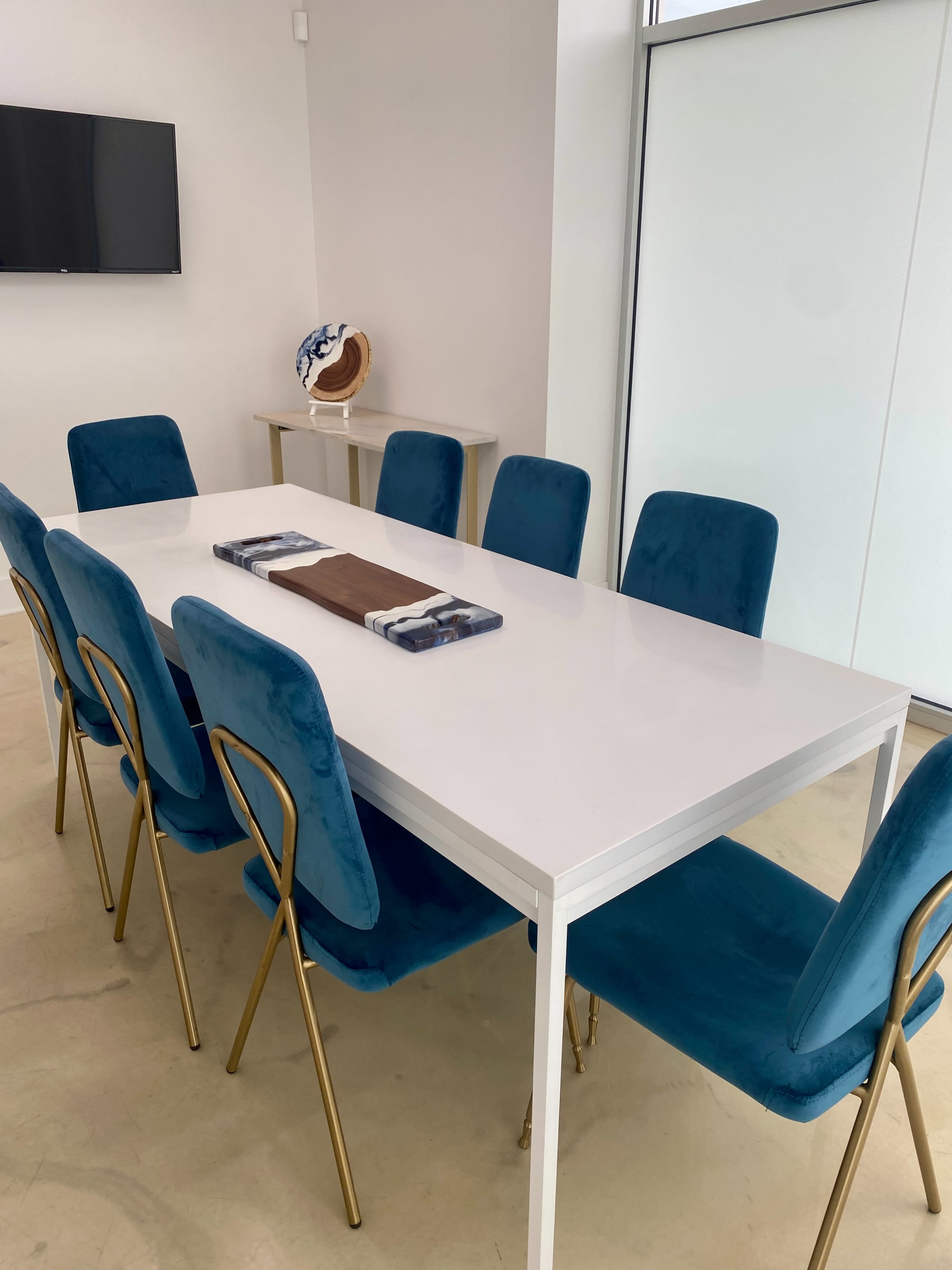 A rectangular white dining table is set with a center runner and surrounded by eight blue upholstered chairs with gold metal legs.