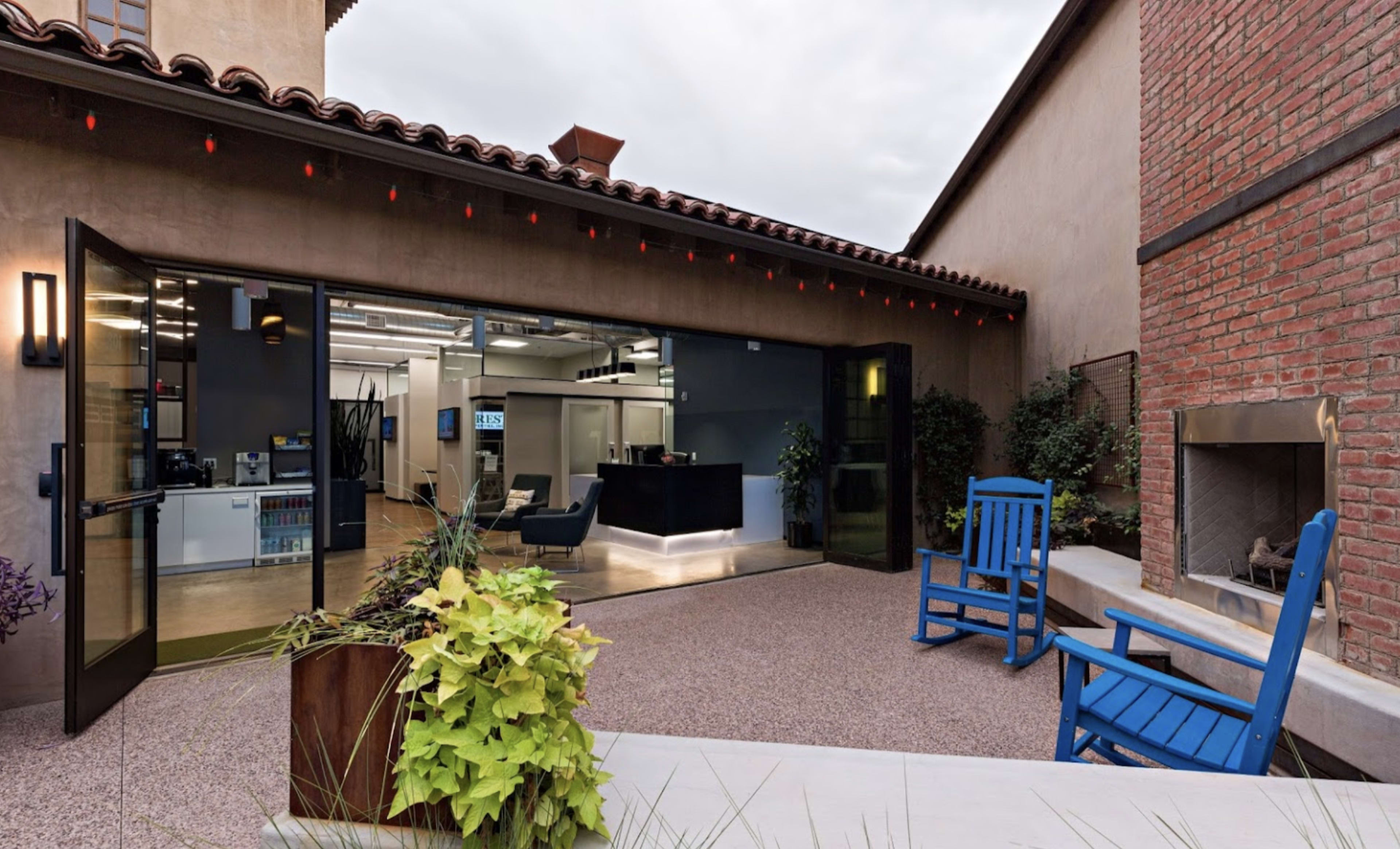 The image shows a modern outdoor patio area with two blue rocking chairs facing a building that has large glass doors opening into a contemporary interior space.