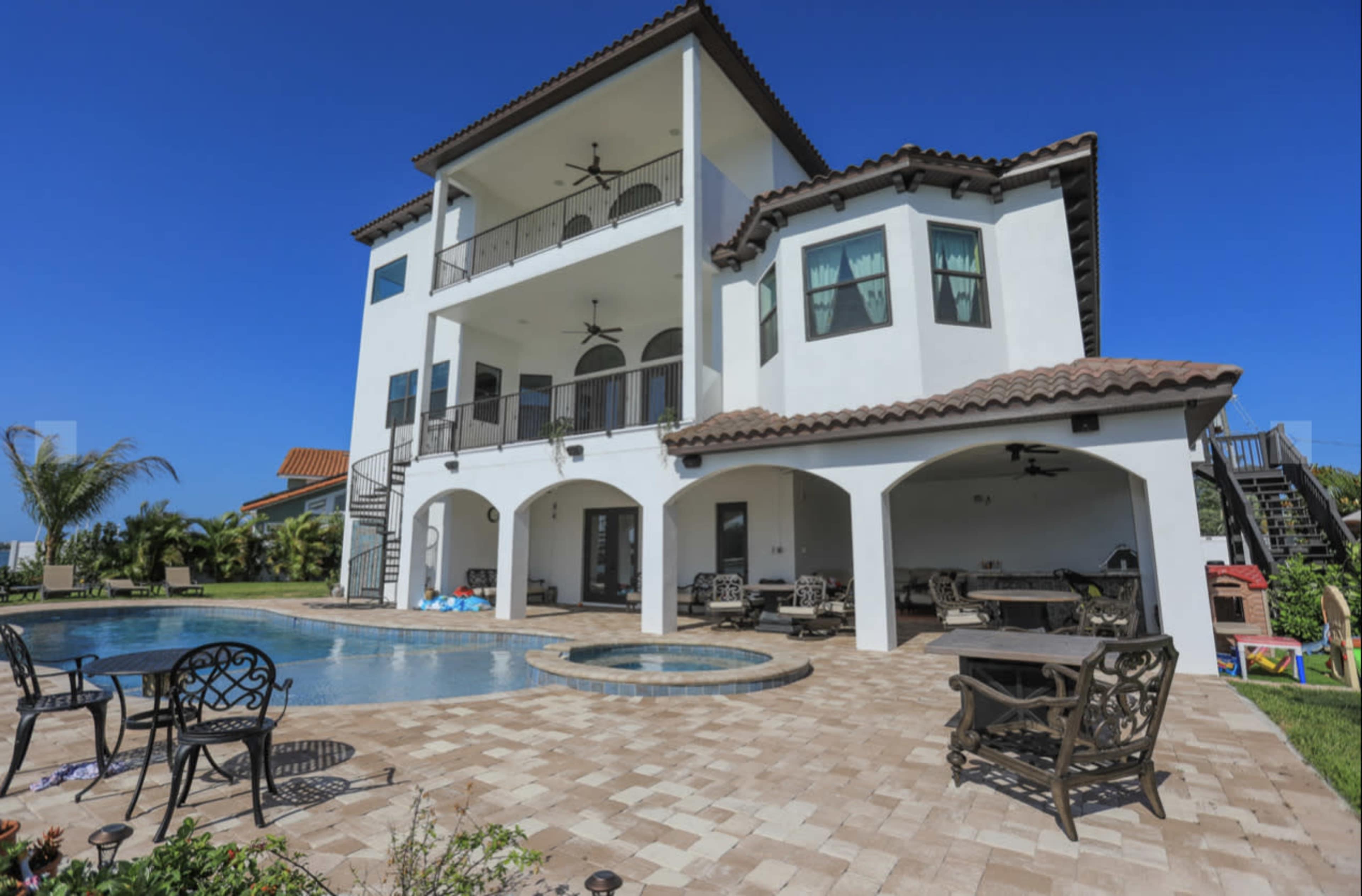 A three-story white house with a tiled roof and a pool is set in a sunny backyard featuring patio furniture and tropical landscaping.