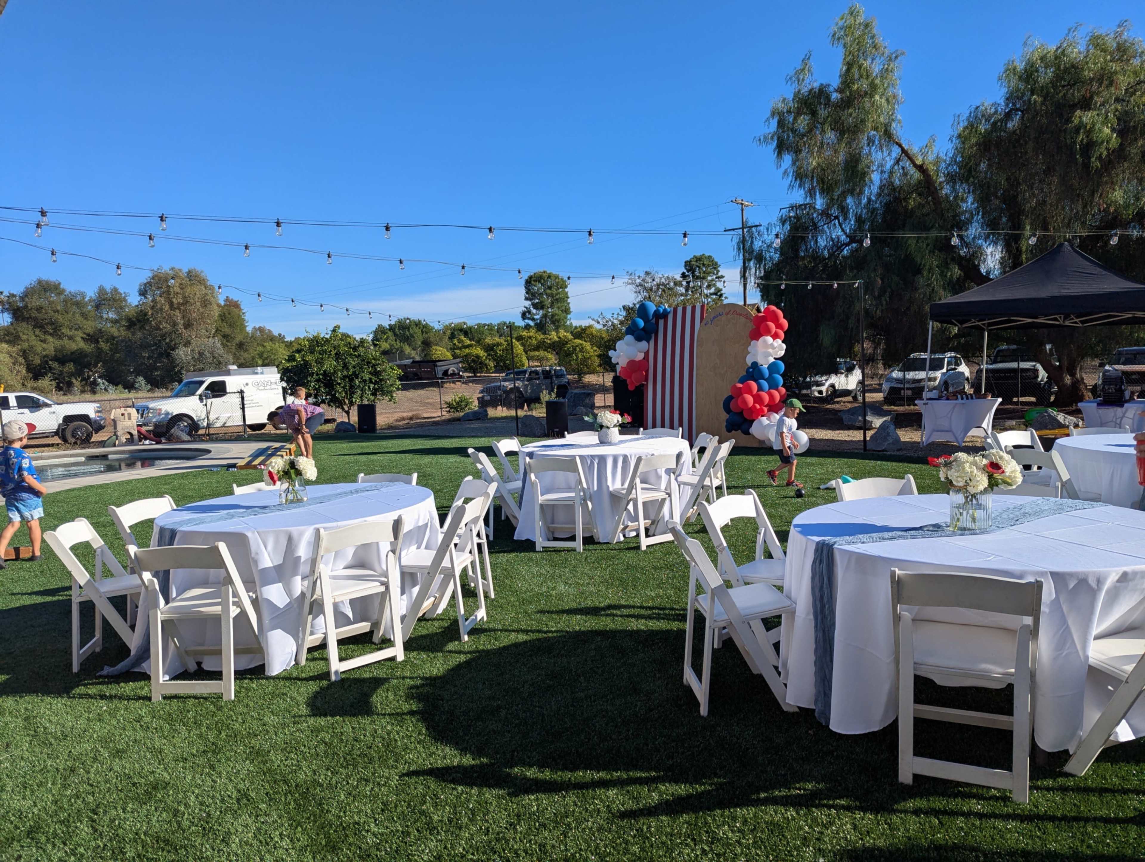 A set of white tables and chairs is arranged on a grassy area, decorated with balloons and a backdrop for an outdoor event.