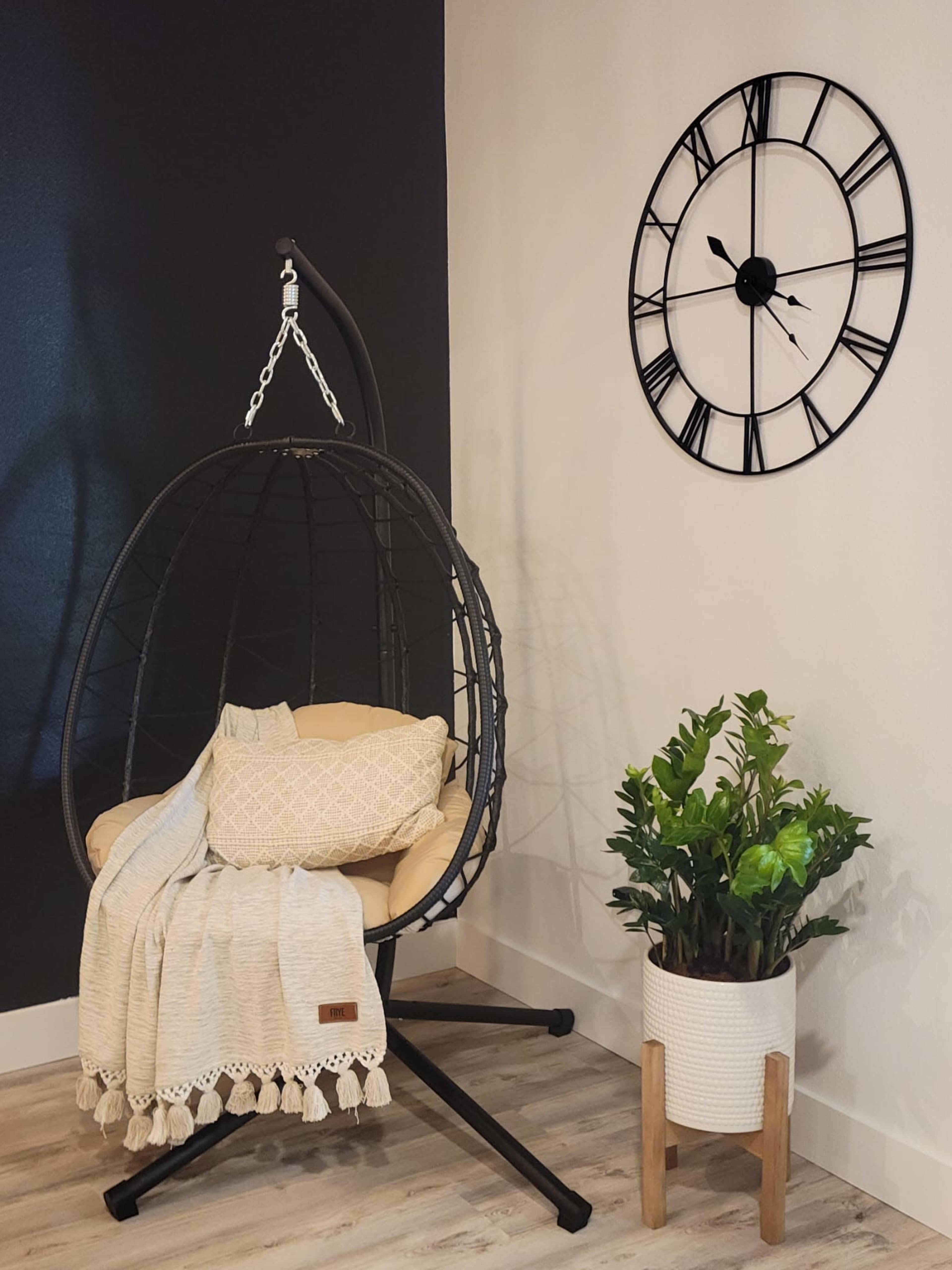 A hanging chair with a cushion and blanket sits next to a modern wall clock and a potted plant.