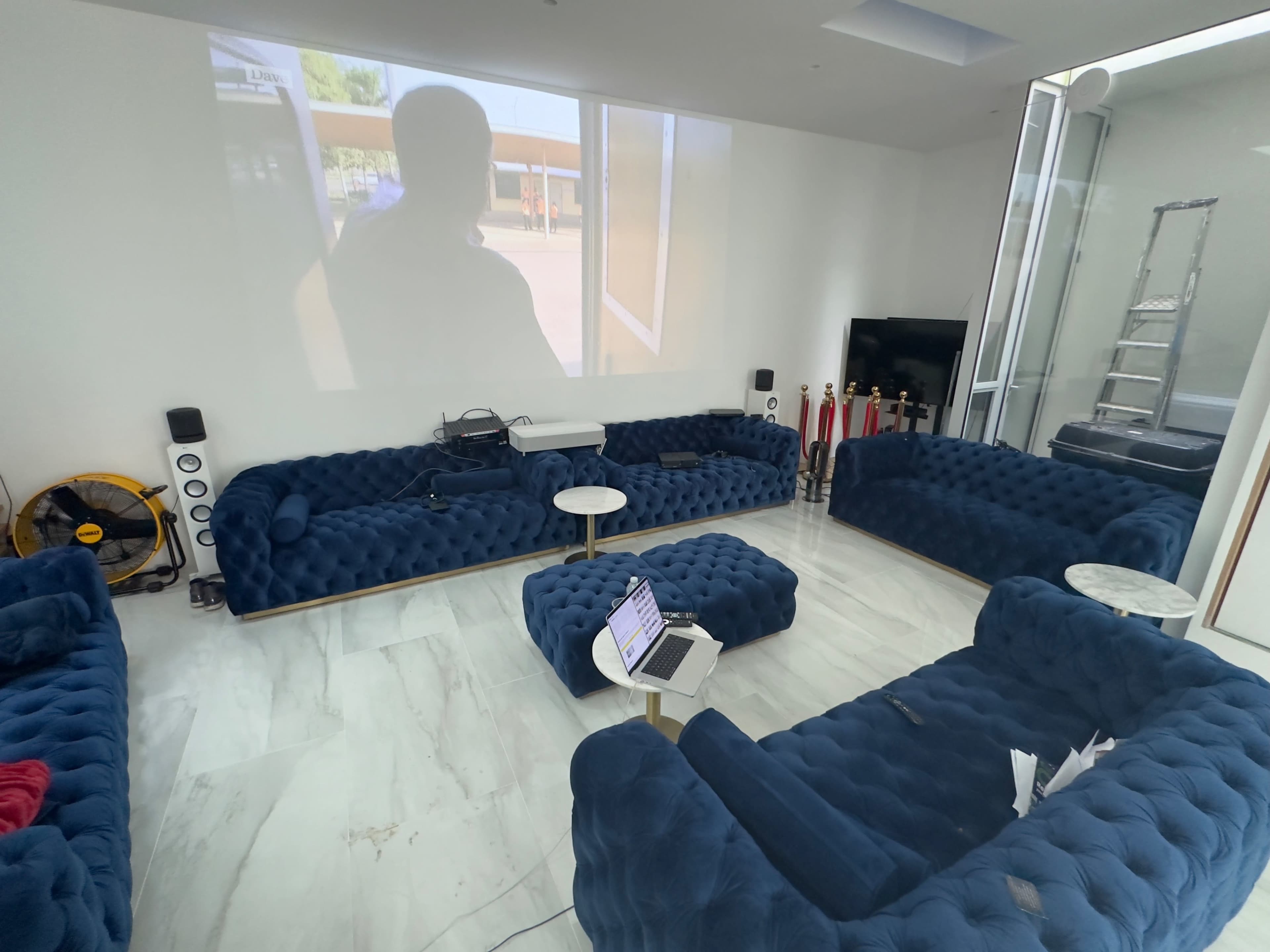 A spacious room with multiple blue tufted sofas arranged around a small white table, featuring a projector screen displaying an outdoor scene.