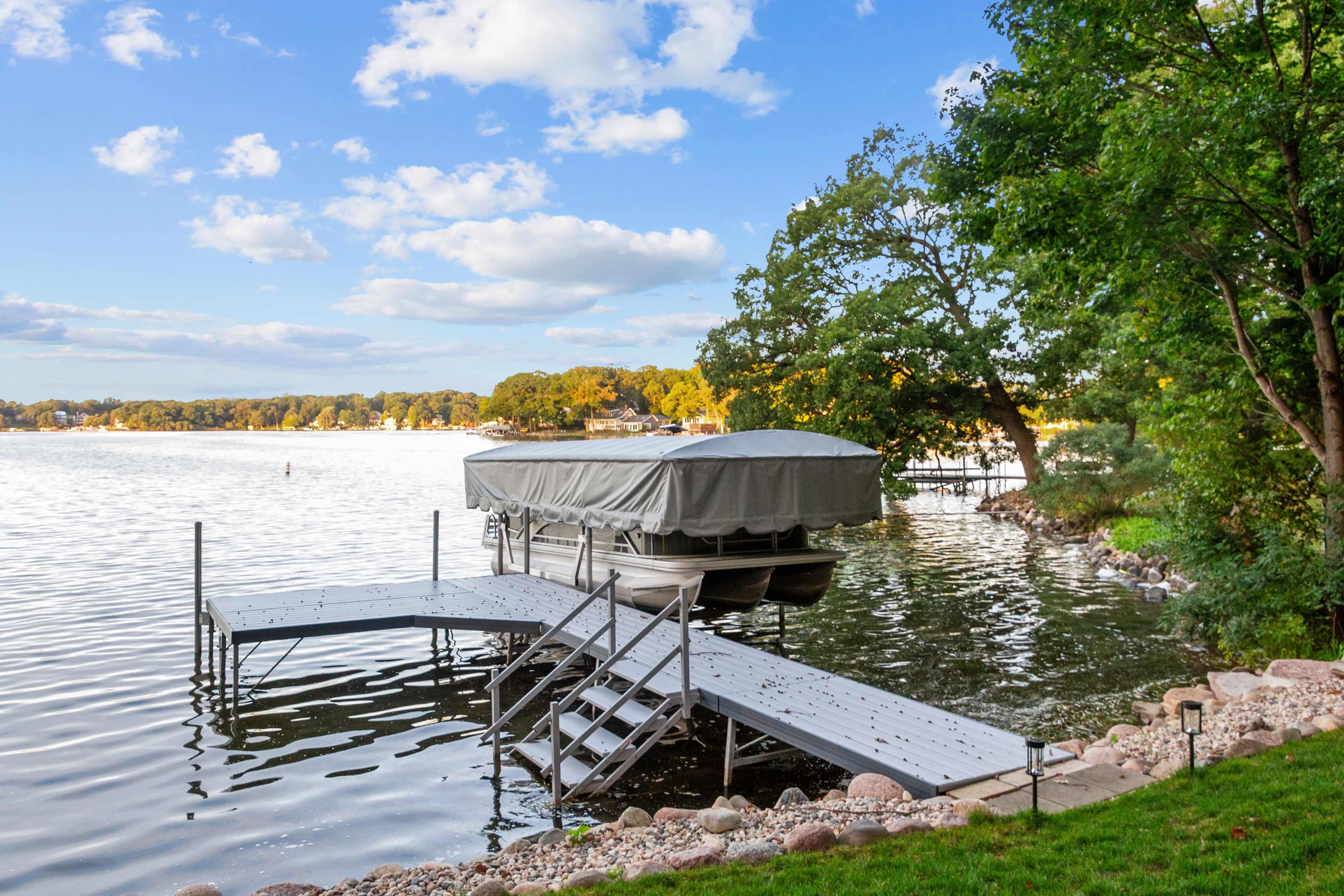 A boat is parked on a covered lift at a dock beside a calm body of water, surrounded by trees and a clear sky.