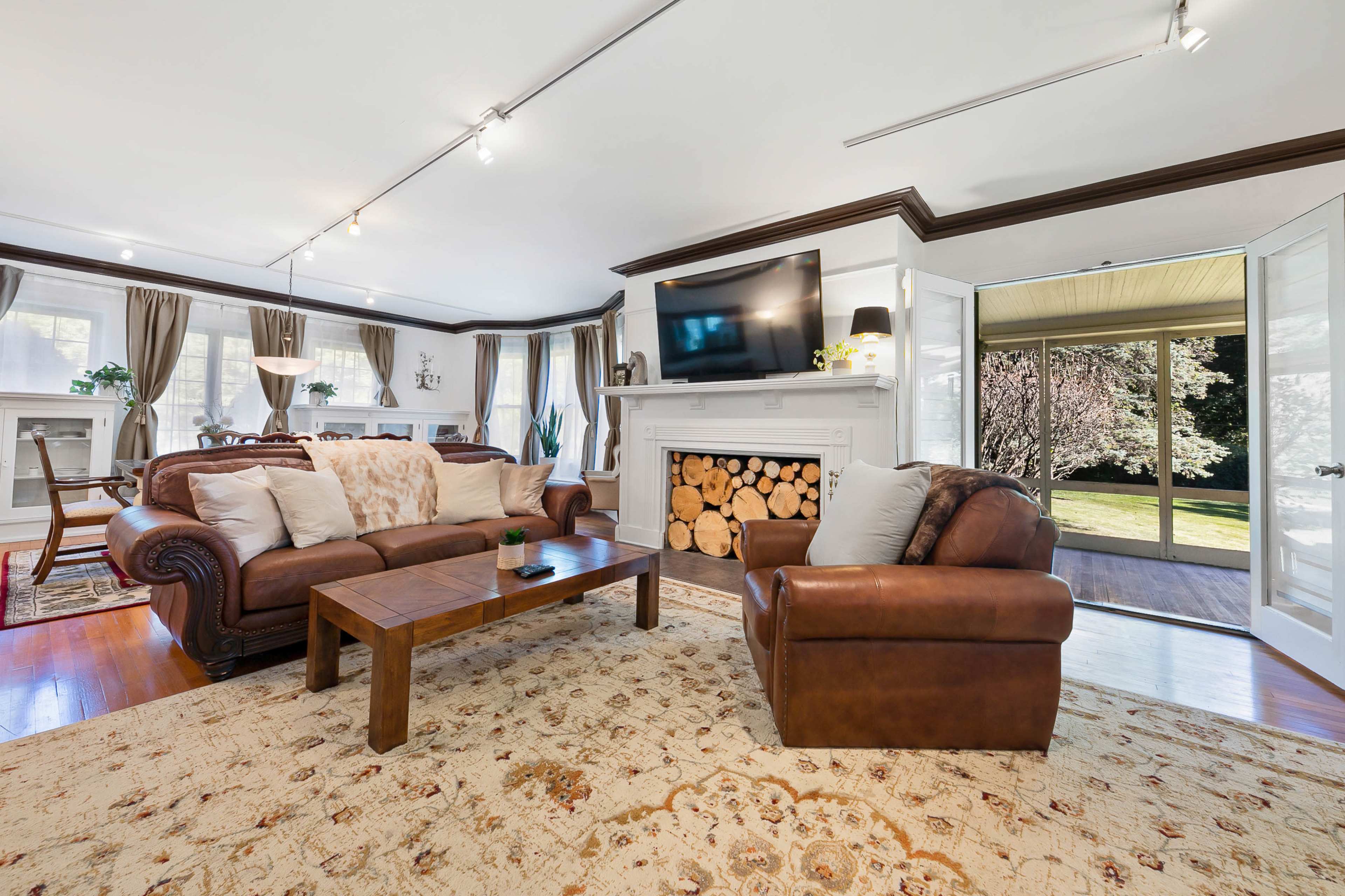 The living room features two leather sofas, a wooden coffee table, and a fireplace with stacked firewood, leading to a sunlit porch with large doors.