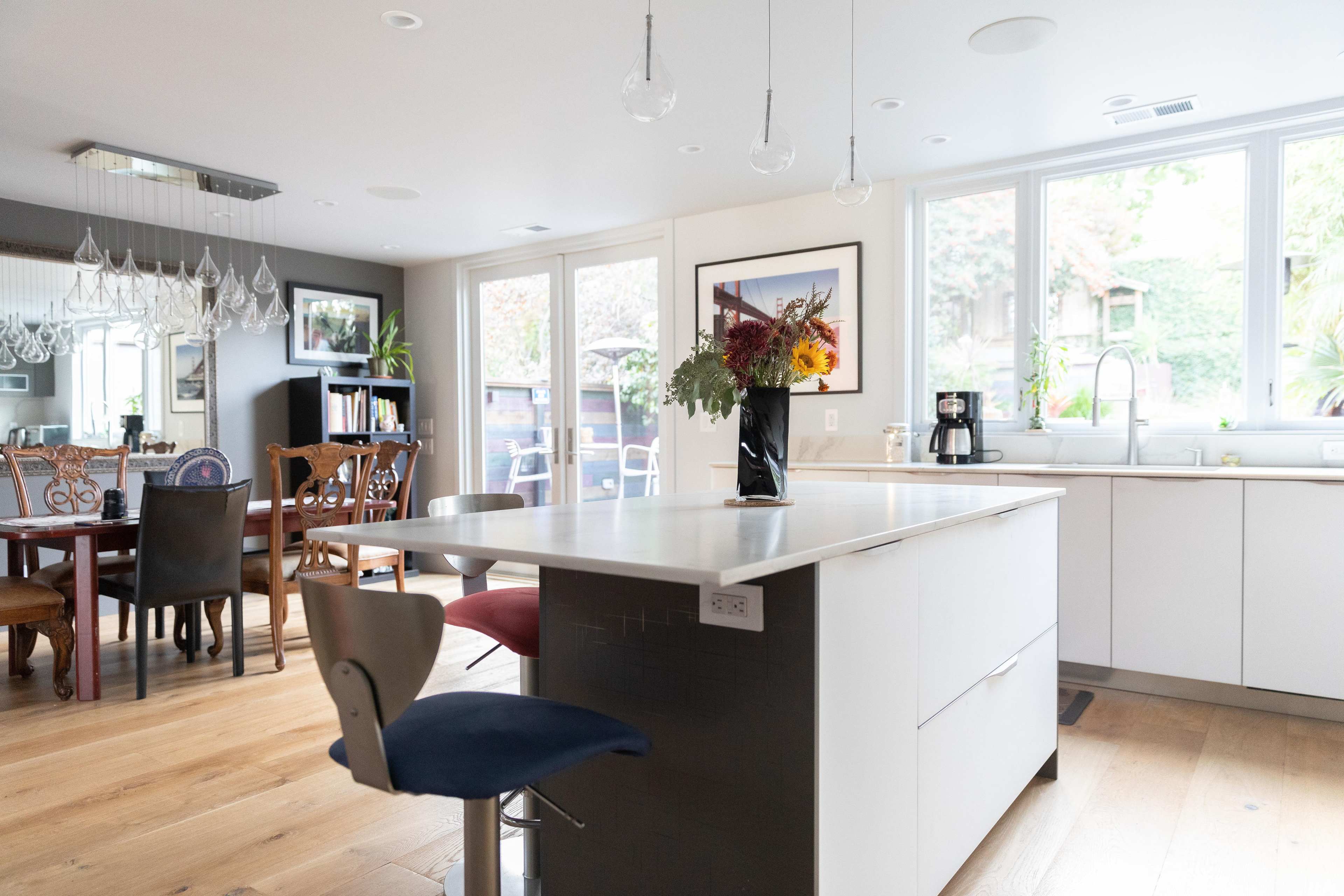 A modern kitchen with a central island, surrounded by large windows that provide natural light, and a dining area seen in the background.