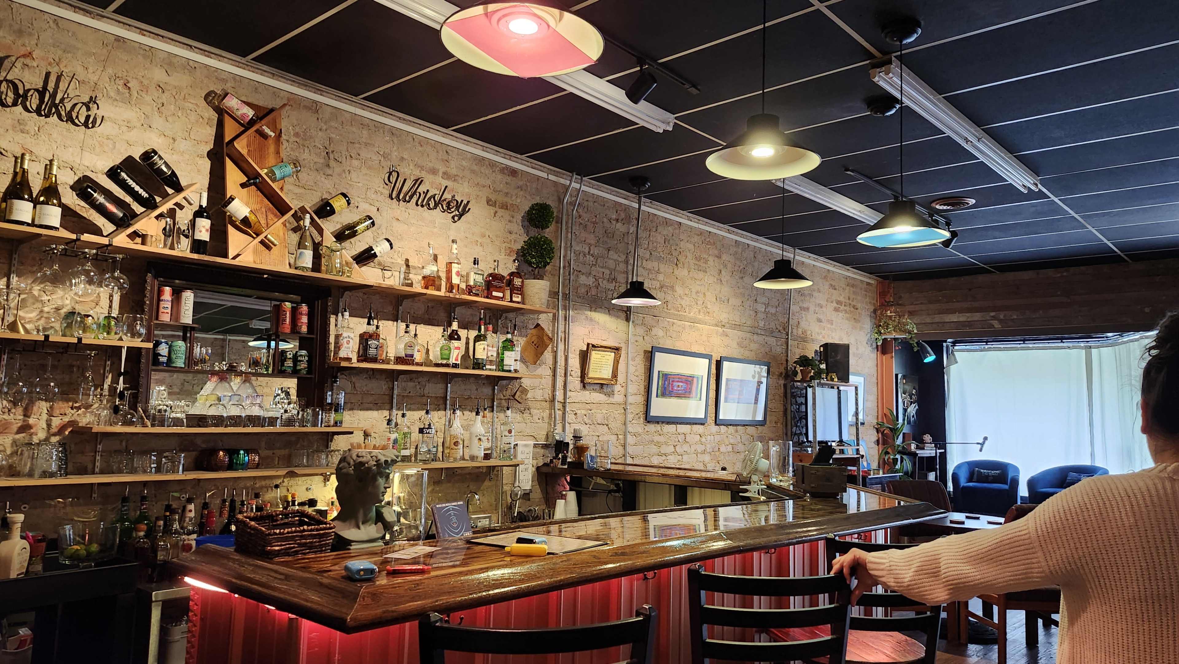 The image shows a bar with a wooden counter, shelves filled with various bottles of alcohol, and colorful pendant lights hanging from the ceiling.