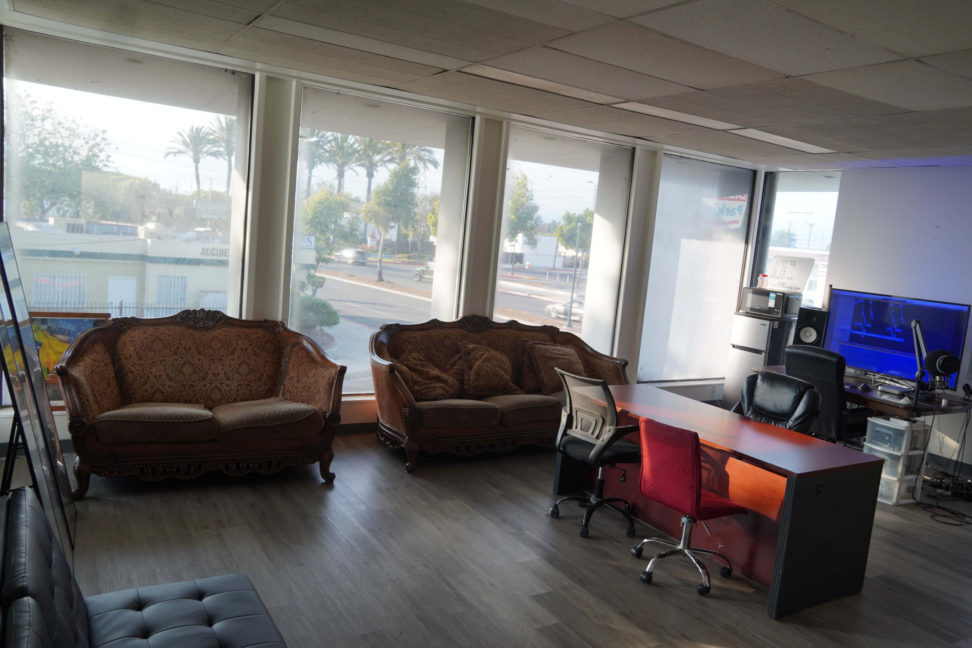 The image shows a spacious office with two ornate couches, a desk with red chairs, and large windows overlooking a street lined with palm trees.