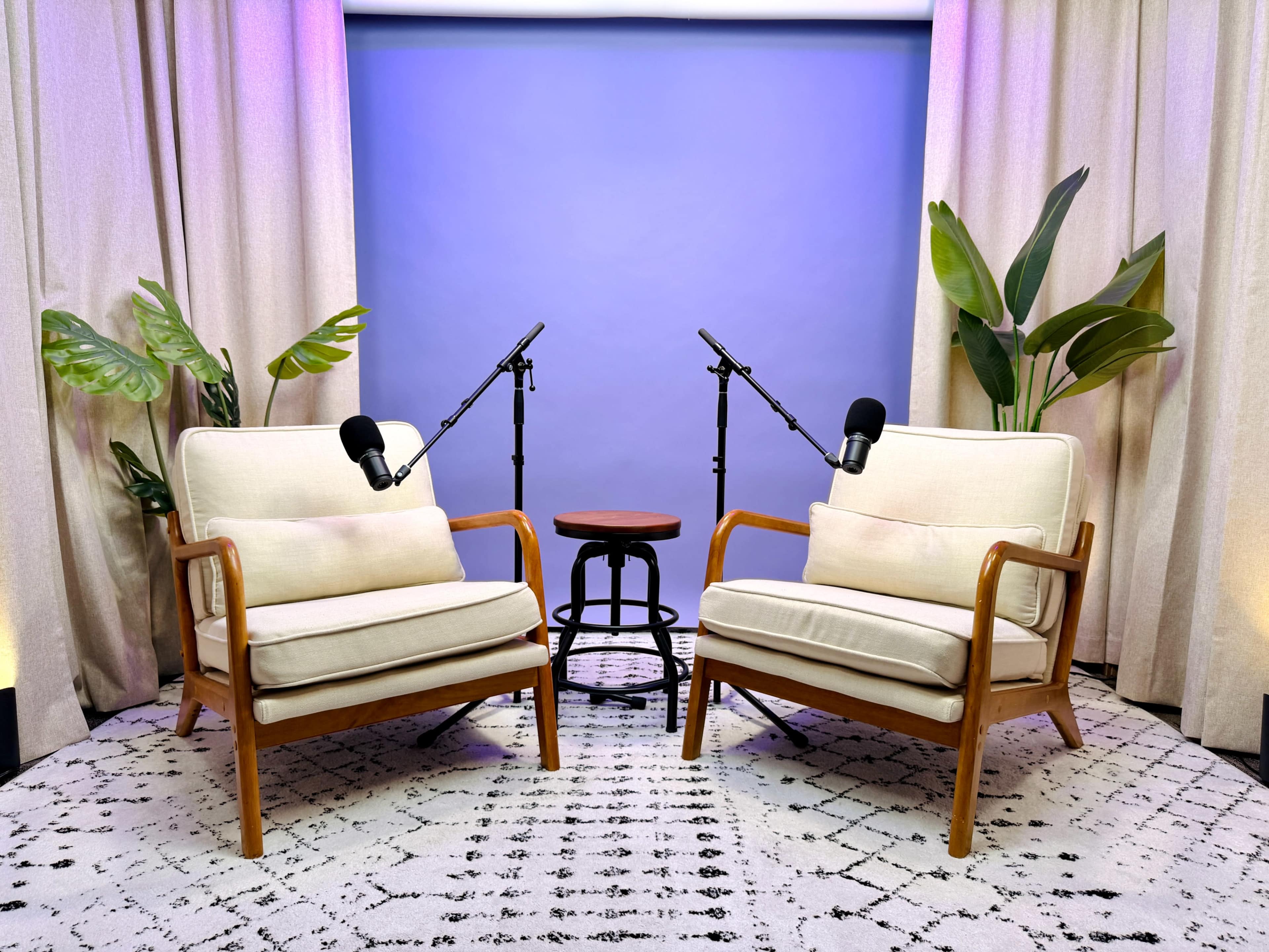 The image shows two cushioned chairs on a patterned rug, positioned opposite each other with a small table and microphones set up between them, surrounded by plants and draped curtains.