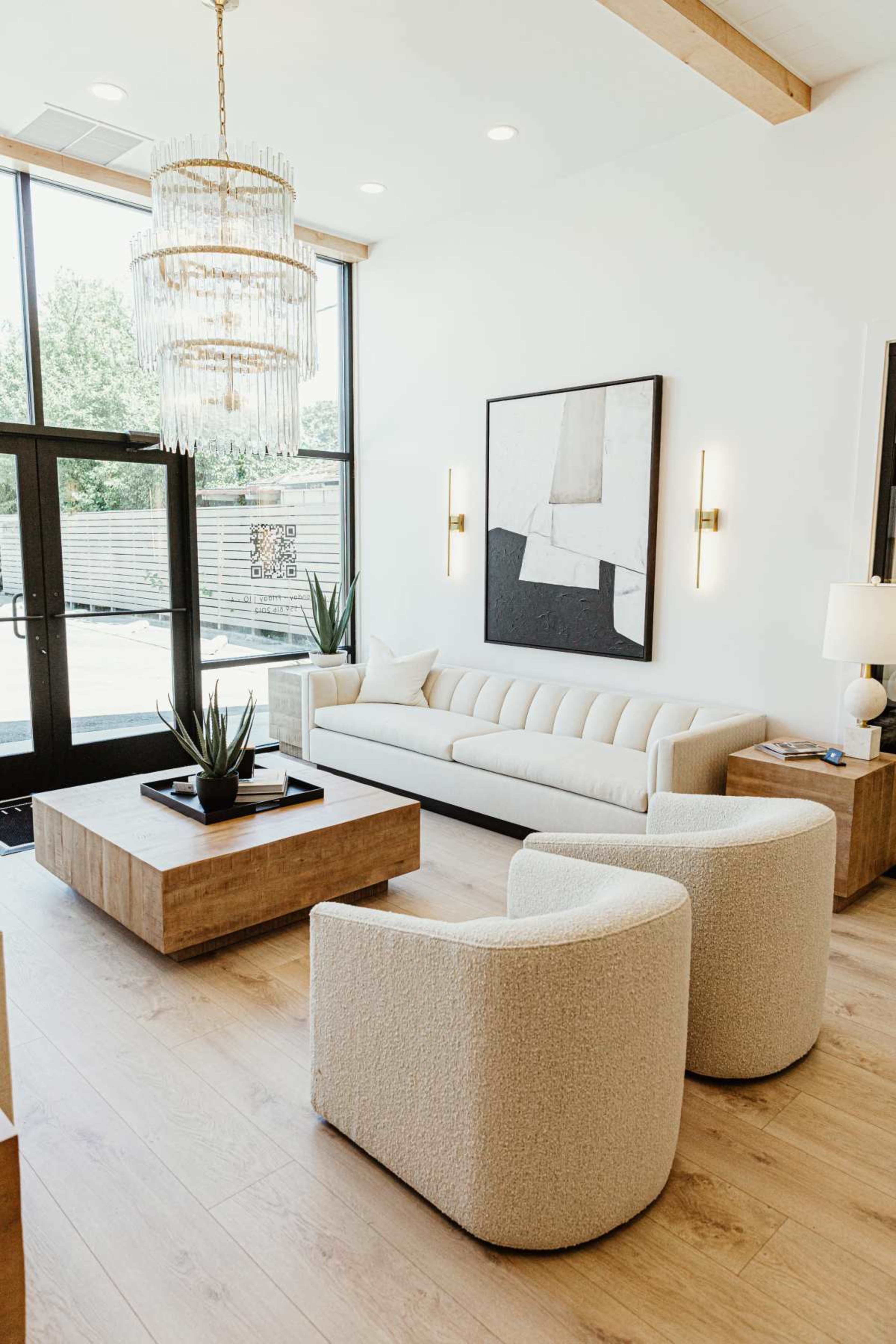 The image shows a modern living room featuring a light-colored sofa, two round chairs, a wooden coffee table, a floor lamp, and a large chandelier, all illuminated by natural light from large windows.
