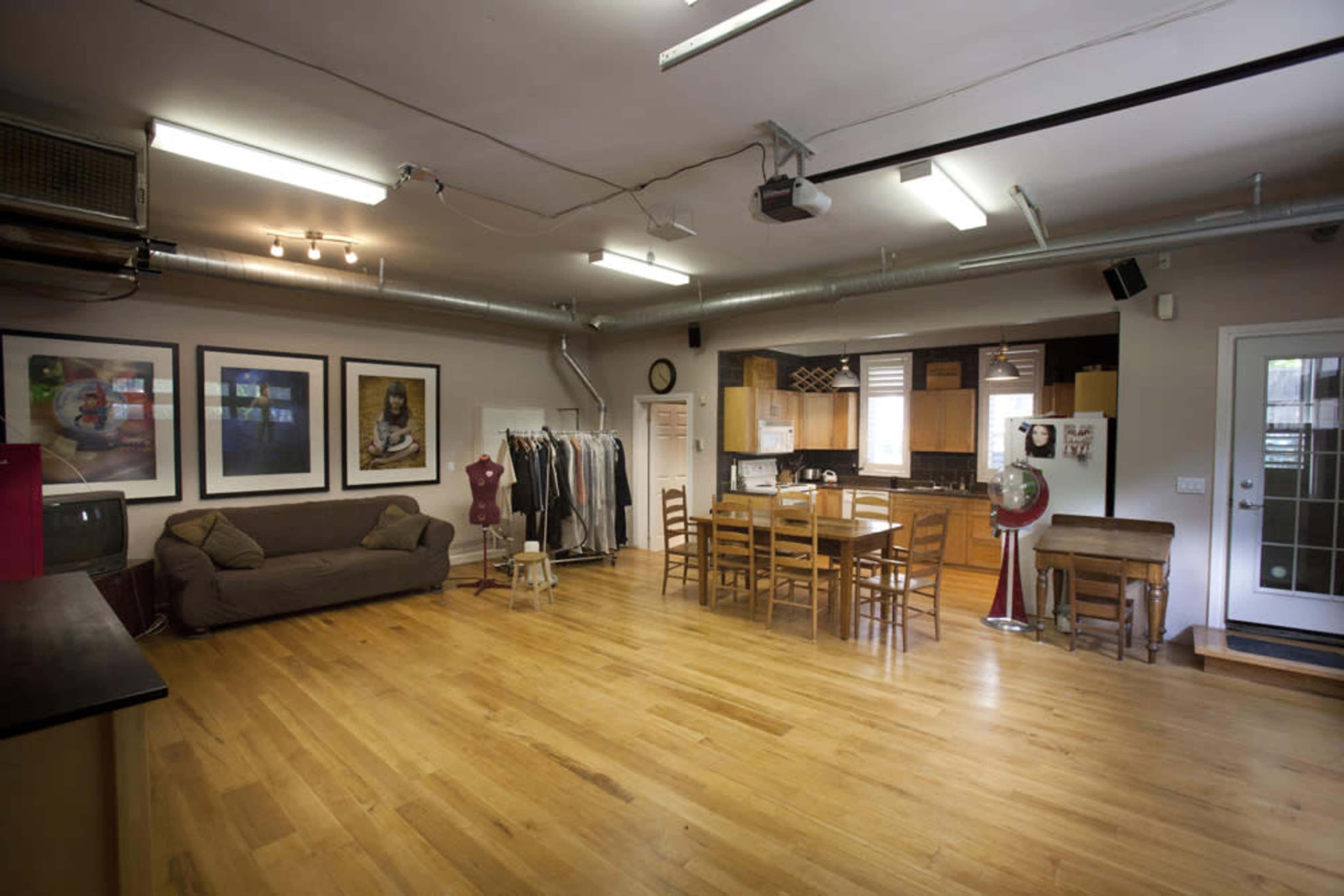 A spacious room with wooden flooring, featuring a sofa, a dining table with chairs, kitchen cabinetry, and a door leading outside.