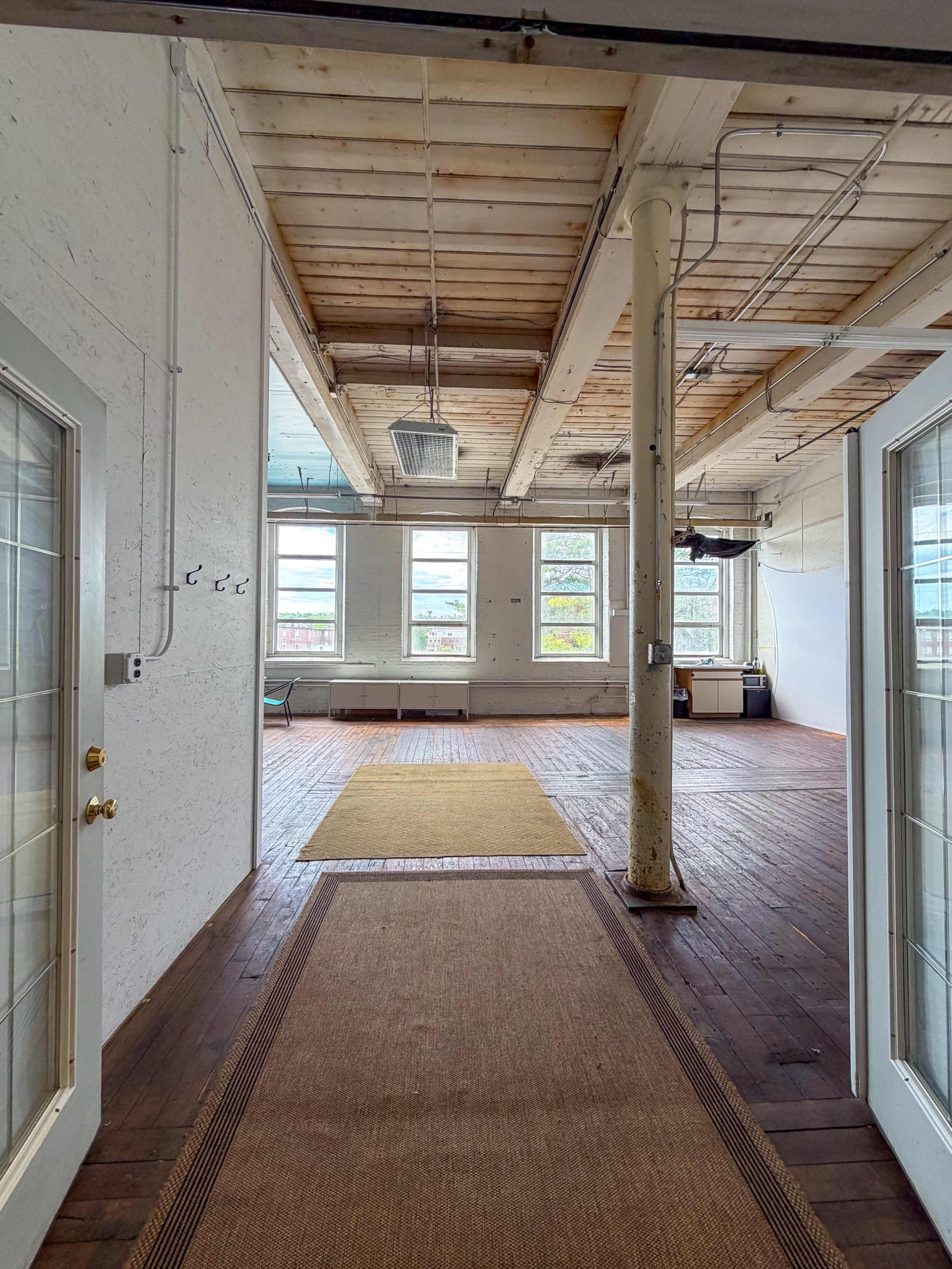 The image shows a spacious, unfinished interior room with wooden floors, large windows, and exposed ceiling beams.