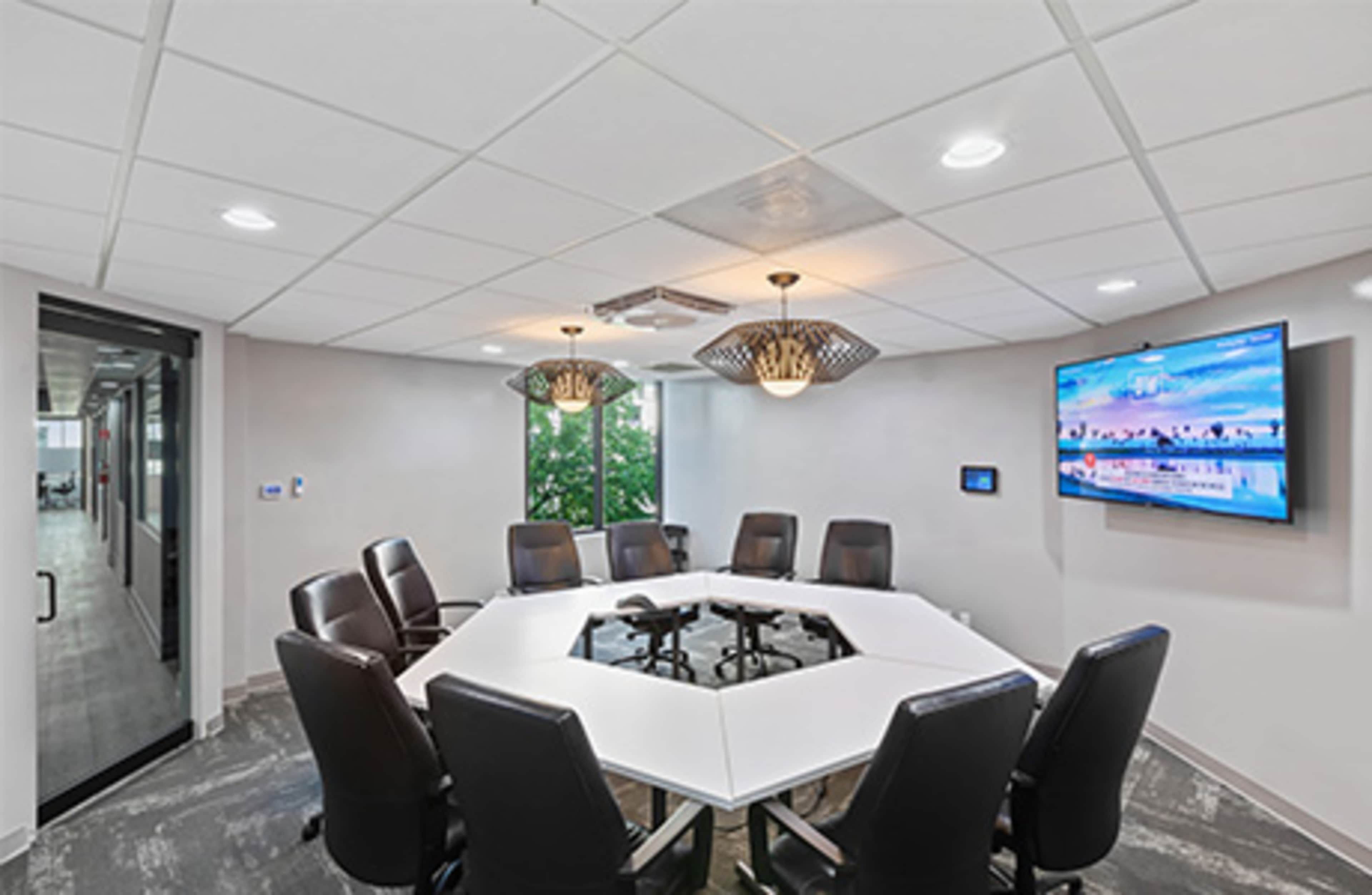 A modern conference room features a large white table surrounded by dark ergonomic chairs, with a wall-mounted screen displaying a peaceful landscape.