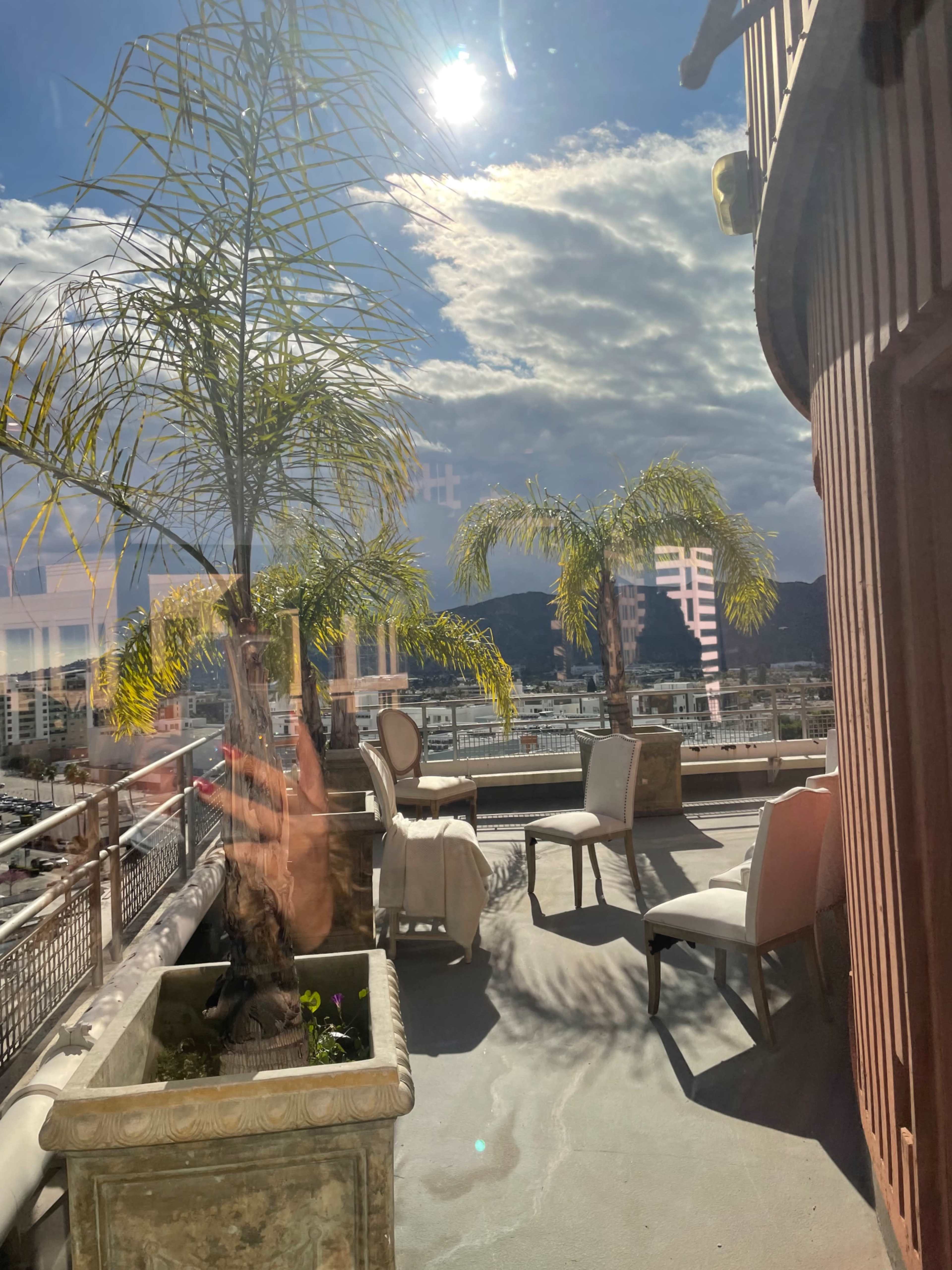 A rooftop terrace features two chairs and palm plants under a bright sky with scattered clouds.