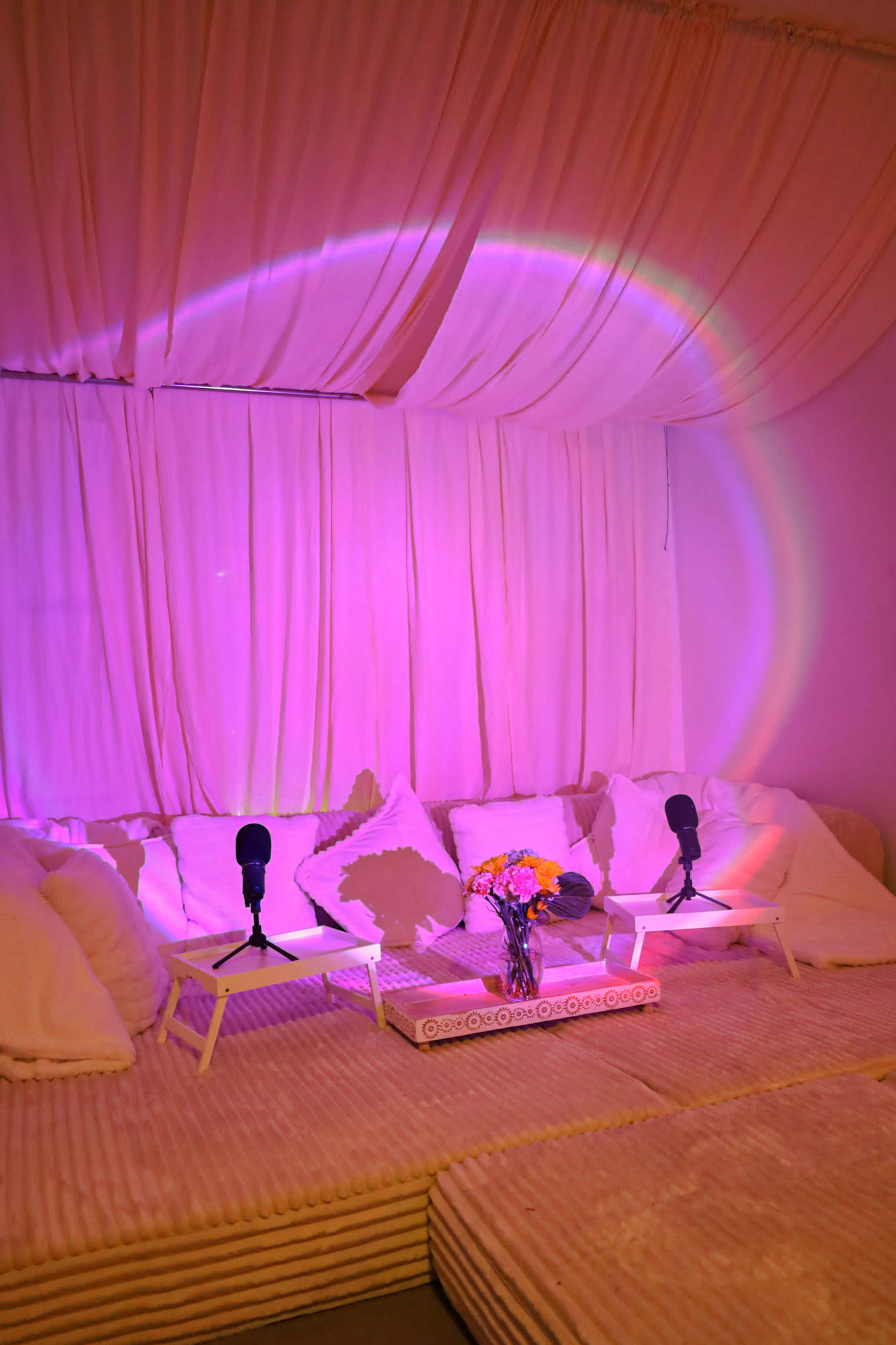 A cozy room features a light-colored couch with white cushions, two microphones on small tables, and soft pink lighting illuminating the space.
