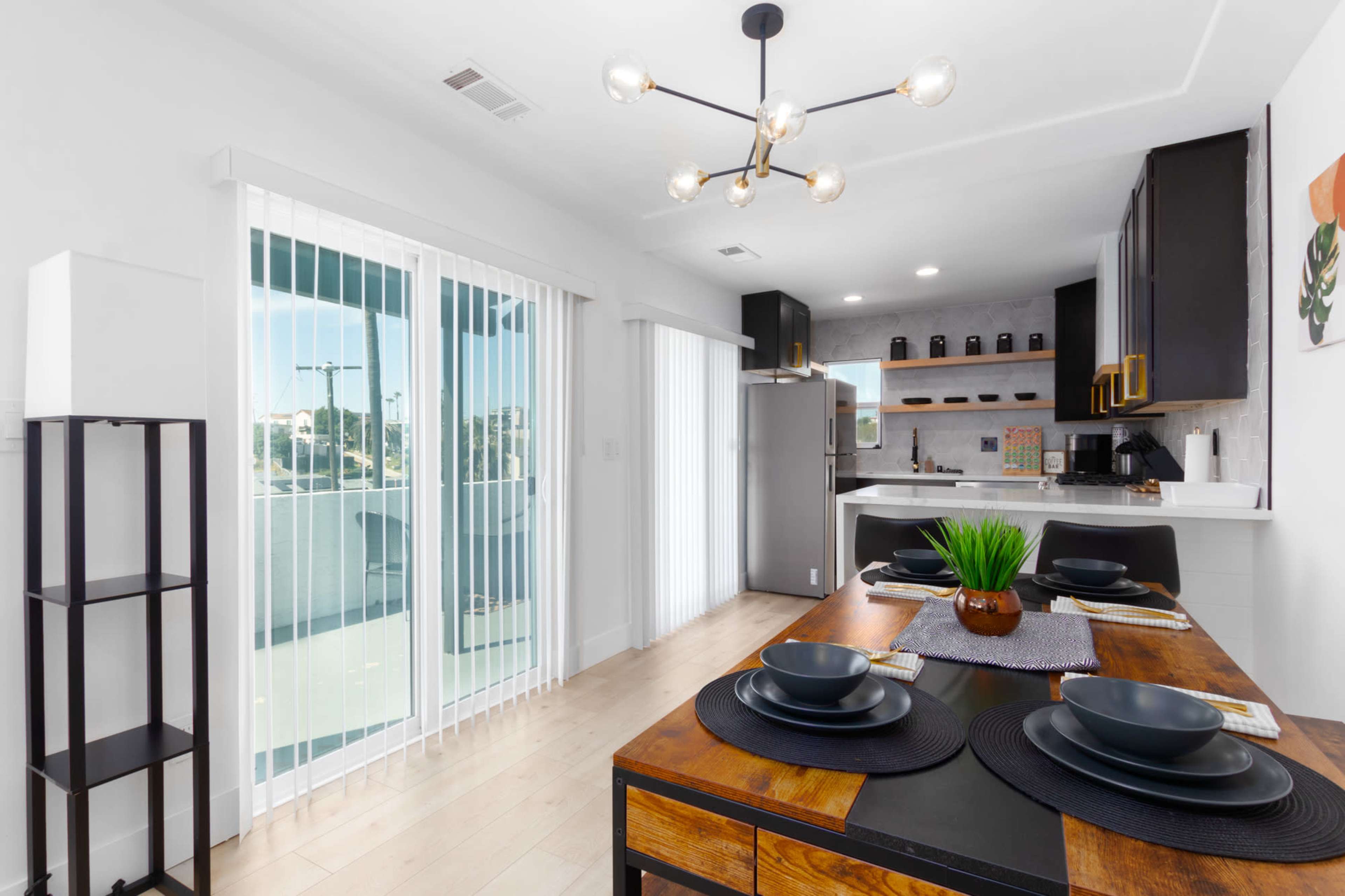 A modern kitchen and dining area feature a wooden table set for four, with a stylish light fixture above and large glass doors leading to a balcony.