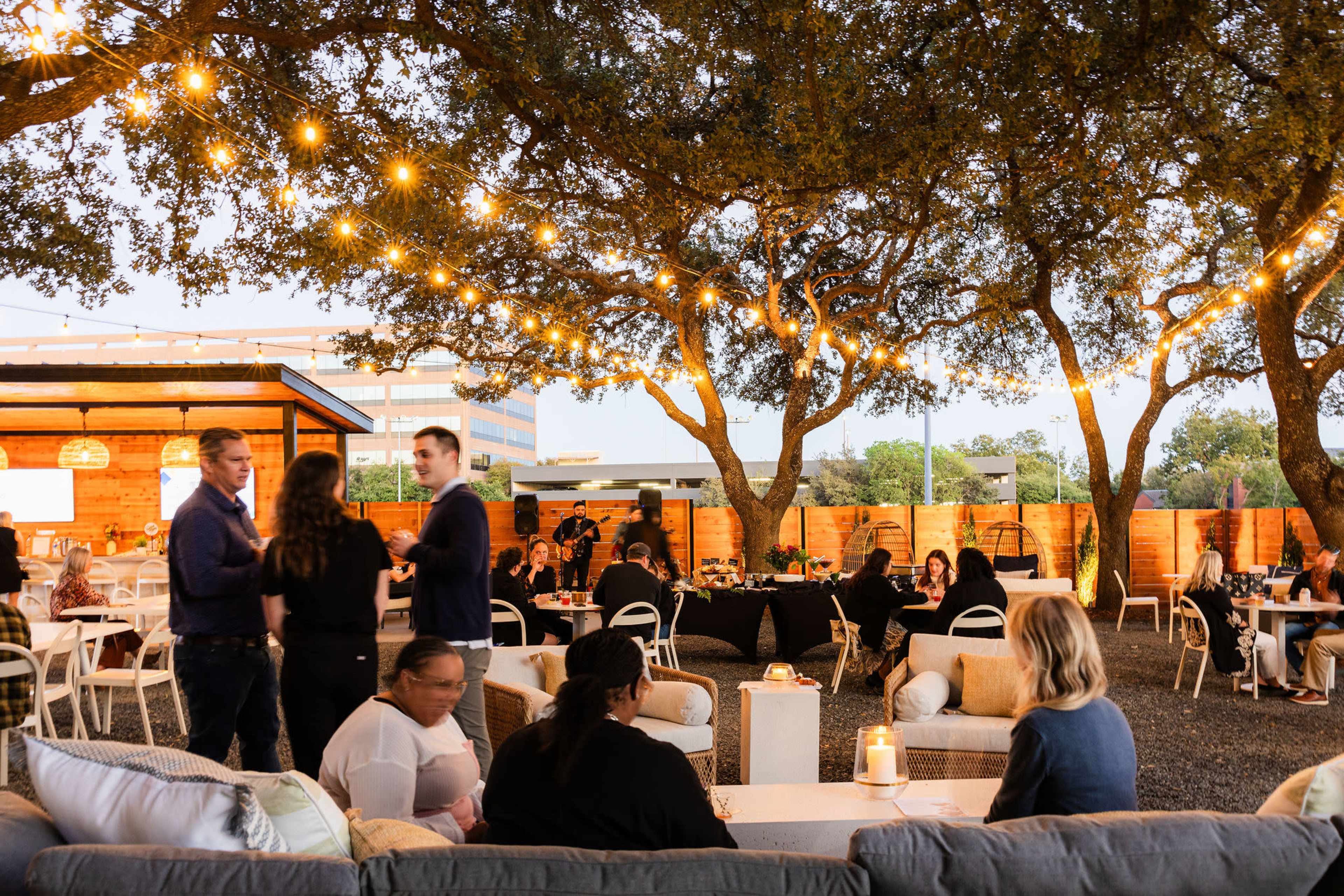 People socialize under string lights hanging from trees in an outdoor event space with seating and tables.