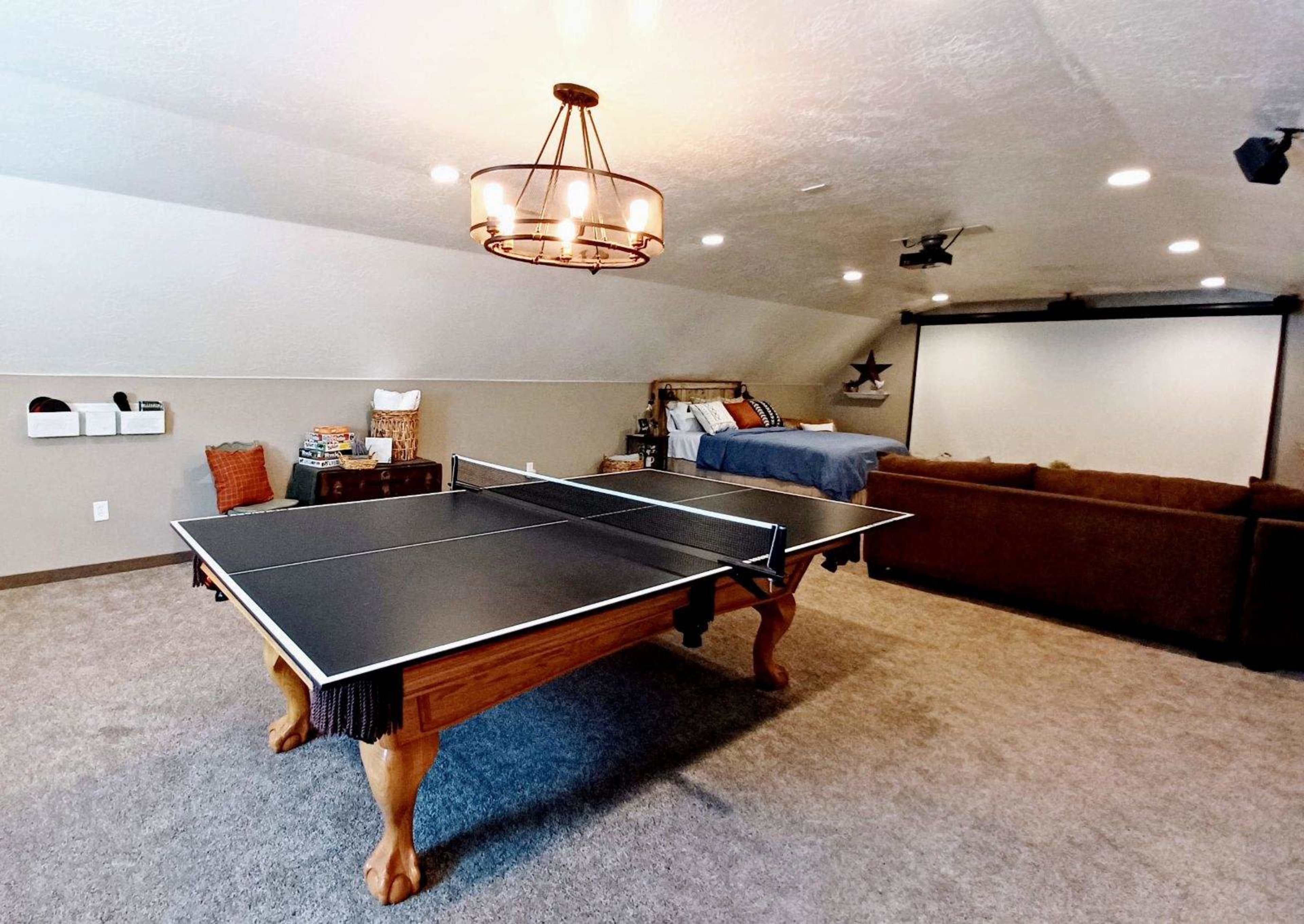 A spacious game room featuring a black ping pong table, a couch, and a bed in a well-lit attic setting.