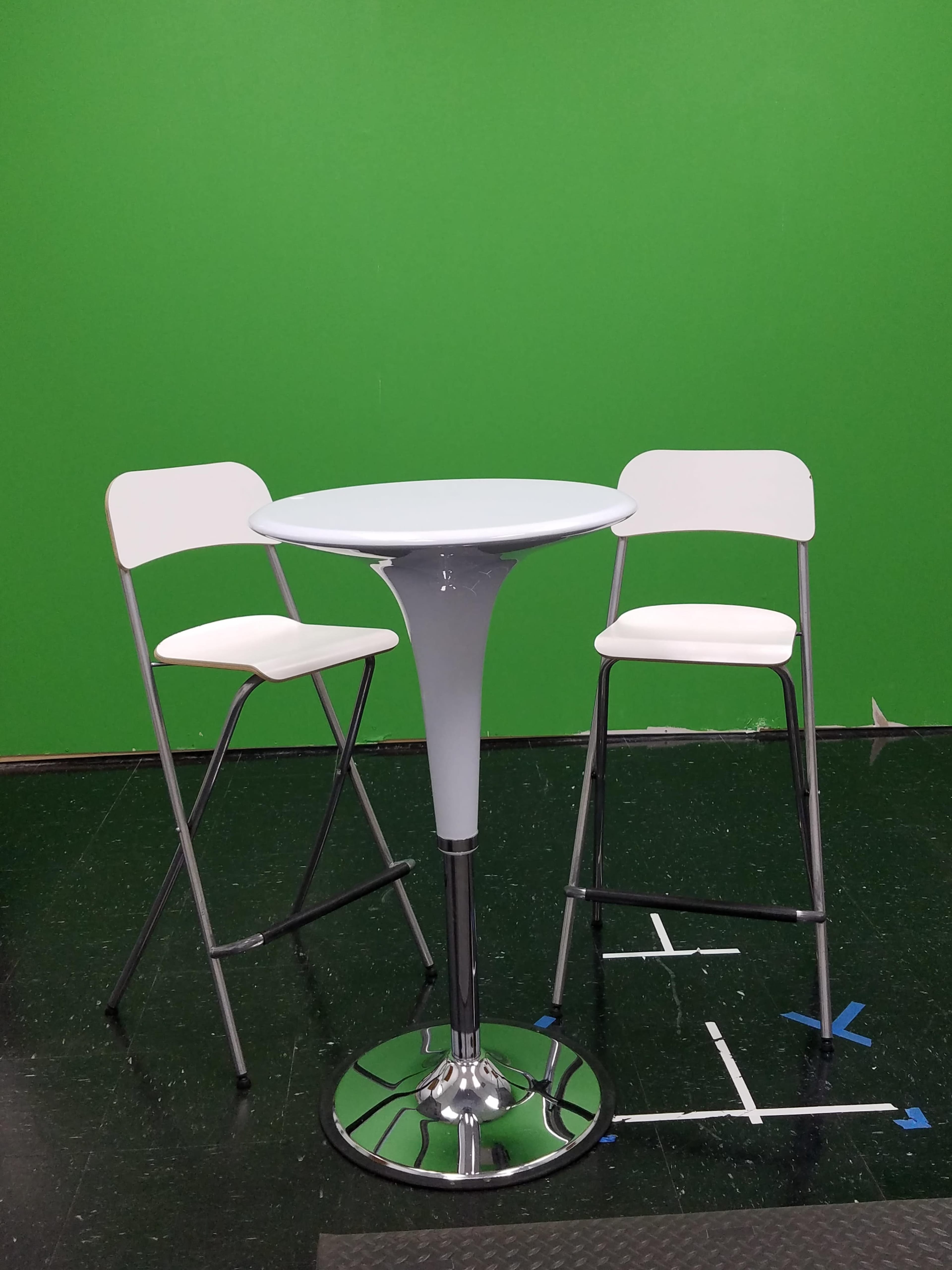 A round table with a white surface is accompanied by two white folding chairs in front of a green backdrop.