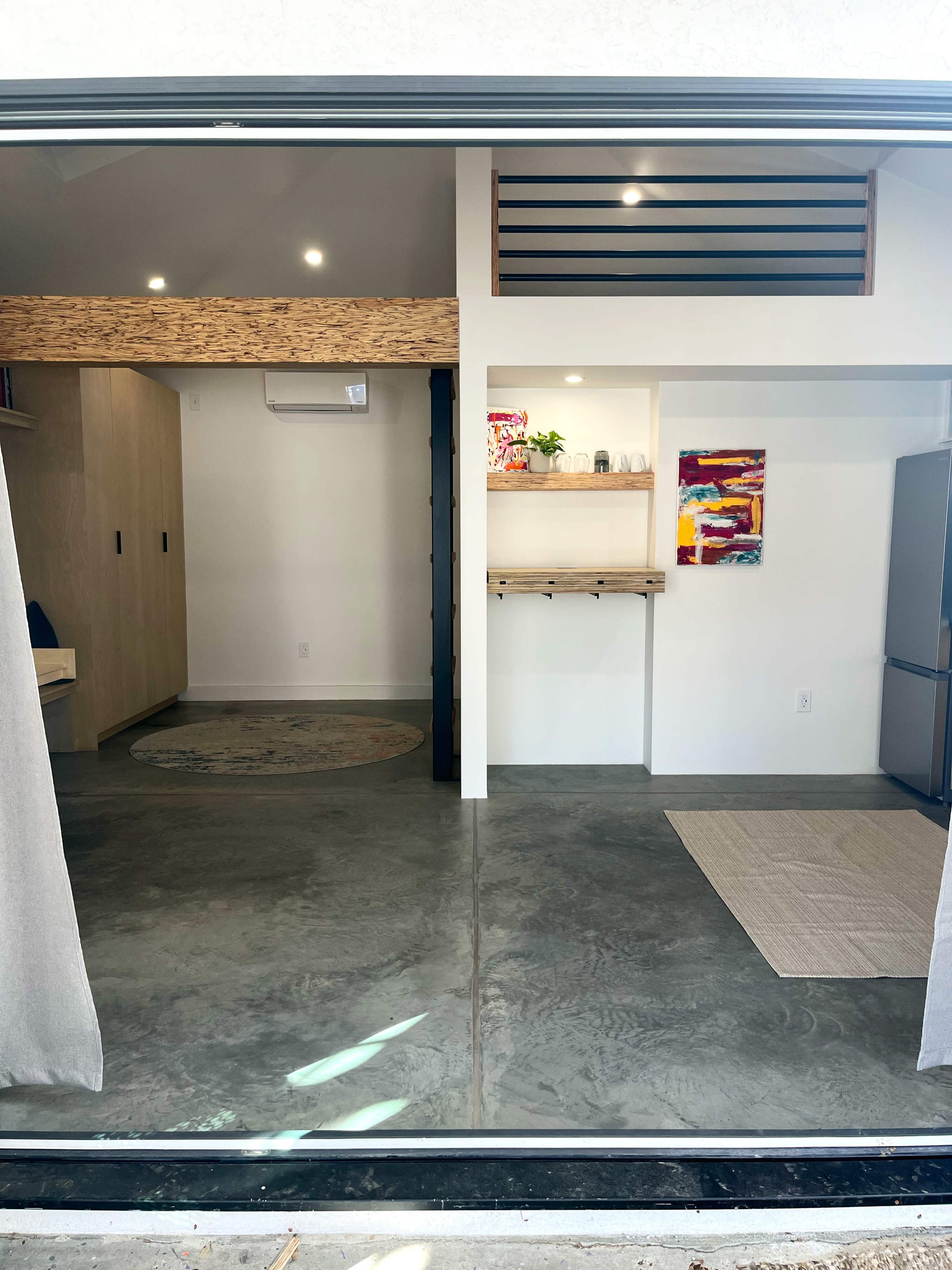 The image shows a modern interior space with concrete flooring, featuring a living area with a circular rug, a kitchenette with wooden shelves, and a bright wall art piece.